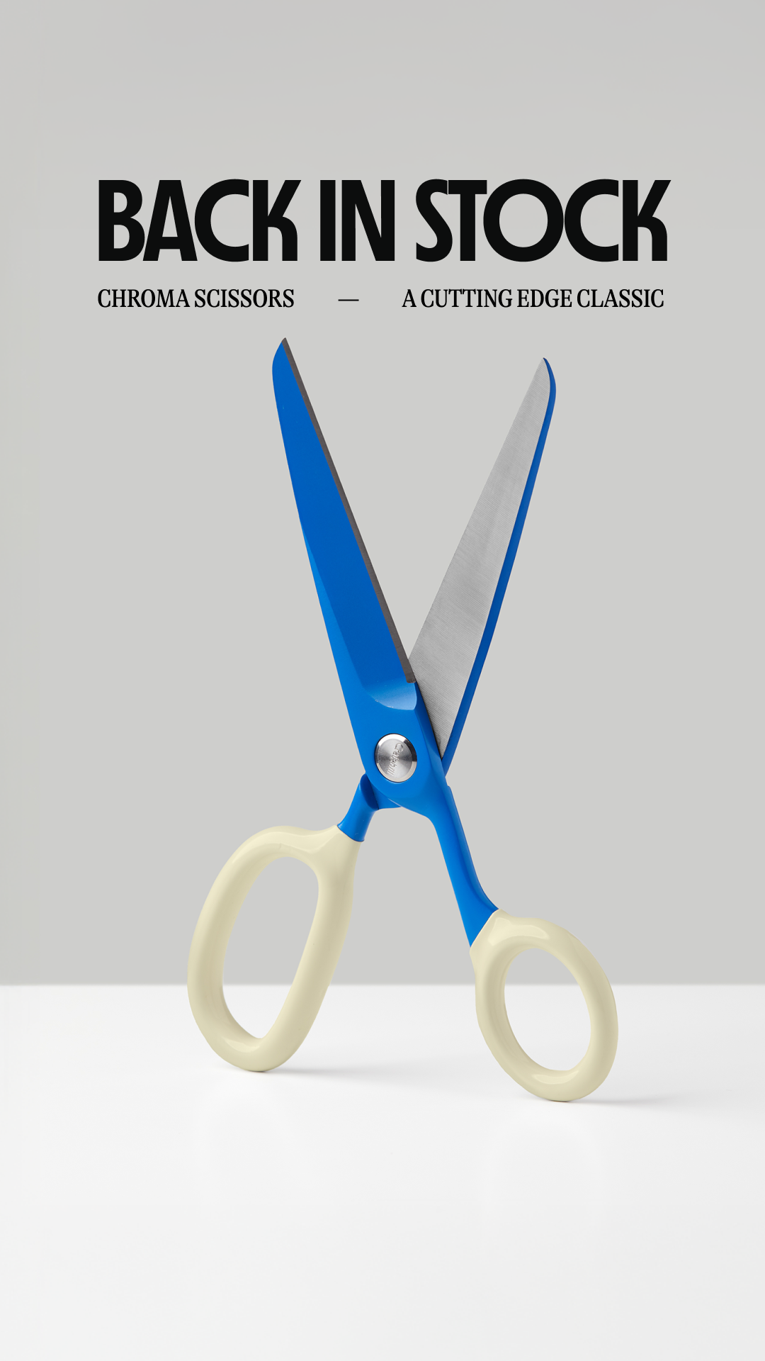 A pair of bright blue and cream-colored scissors are displayed prominently against a plain grey background. The scissors are open, with their blades facing upwards. Above the scissors, large, bold black text reads "BACK IN STOCK". Below this, smaller text states "CHROMA SCISSORS" and "A CUTTING EDGE CLASSIC". The overall impression is a clean, product-focused advertisement highlighting the availability of the scissors.