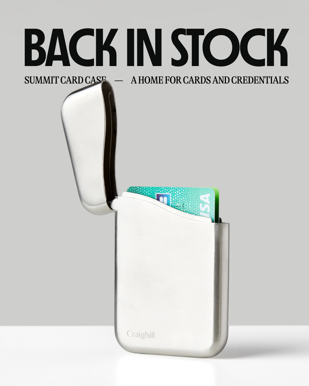 A clean, studio shot of a silver metal card case with its lid open, revealing a Visa card inside. The text "BACK IN STOCK" is prominently displayed in large, bold, black sans-serif font at the top of the image. Below it, in smaller black text, reads "SUMMIT CARD CASE — A HOME FOR CARDS AND CREDENTIALS". The brand name "Craighill" is subtly visible in grey on the front of the card case. The background is a plain, light grey gradient.