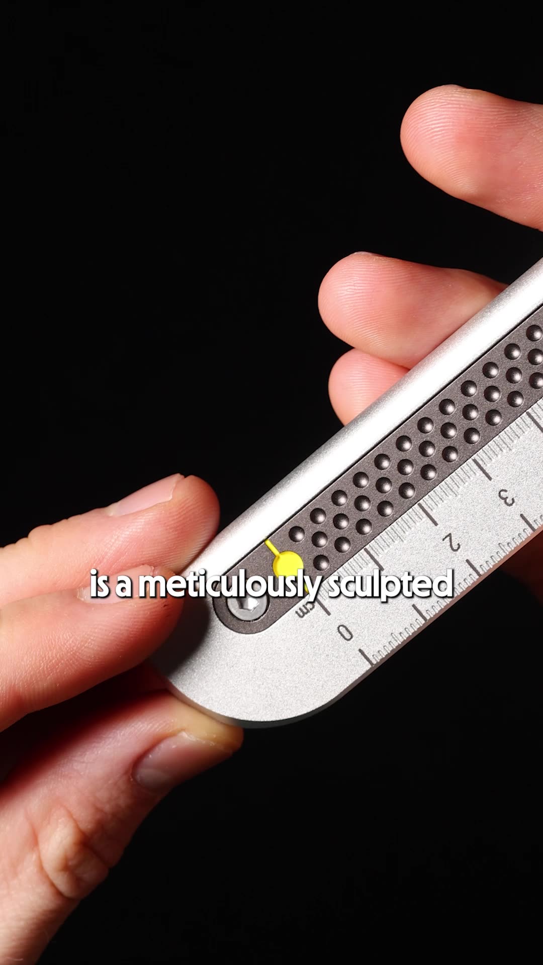 A close-up shot focuses on a person's hands holding a metallic ruler with measurement markings. The ruler has a textured grip area with small circular indentations and a yellow marker. White text overlaid on the image reads "is a meticulously sculpted". The background is dark and out of focus, drawing attention to the product and the text. The creative highlights a specific feature of the ruler.