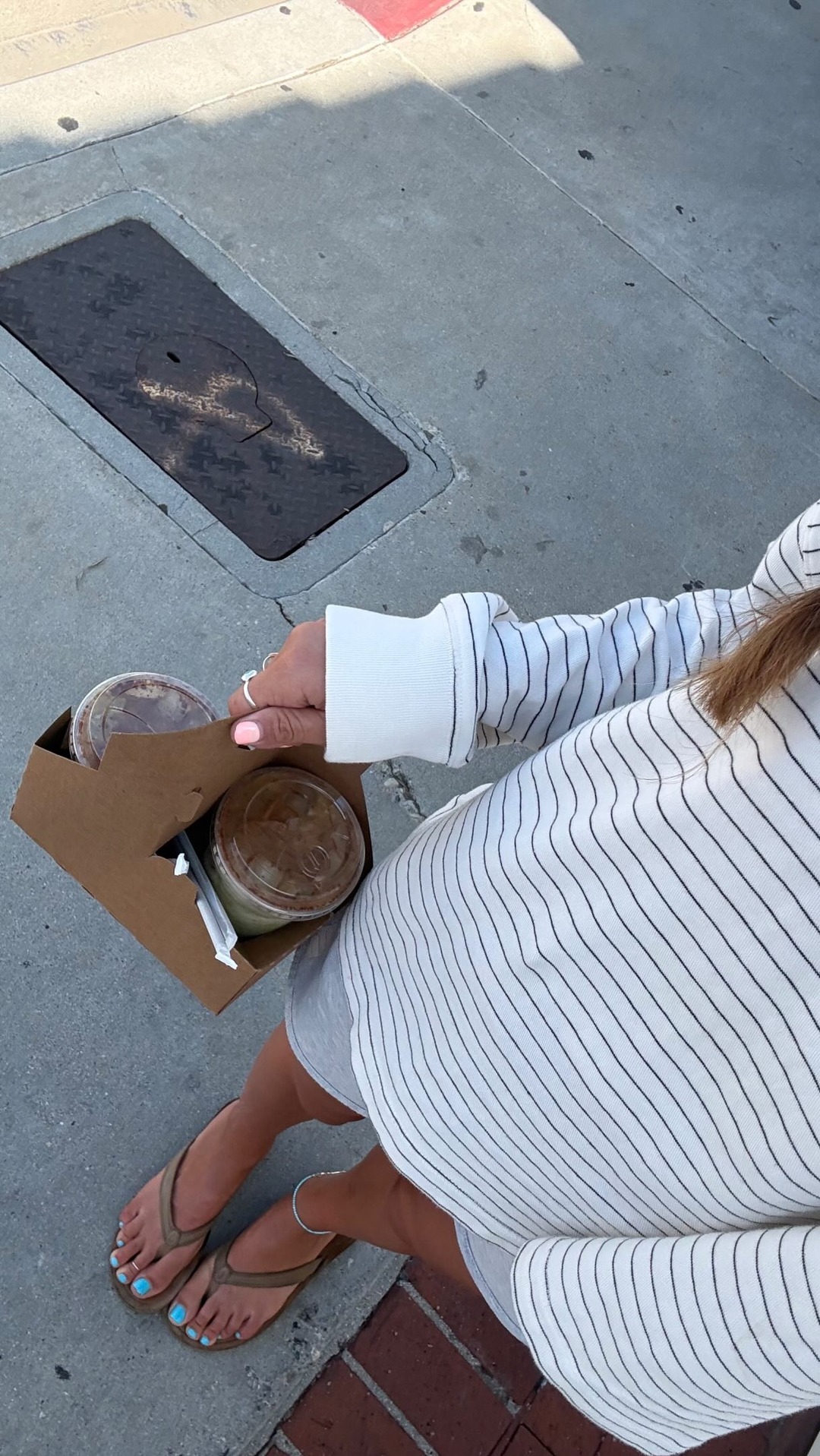 A person's hand, adorned with a ring and light pink nail polish, holds a brown cardboard carrier containing two to-go cups of iced beverages. The person is wearing a white and black striped long-sleeved shirt and grey shorts, with their feet in brown flip-flops visible at the bottom. The scene is set on a concrete sidewalk with a metal grate in the background. Text overlays the image, announcing 'Costco haul, snack edition!' and mentioning Chomps turkey chomplings.