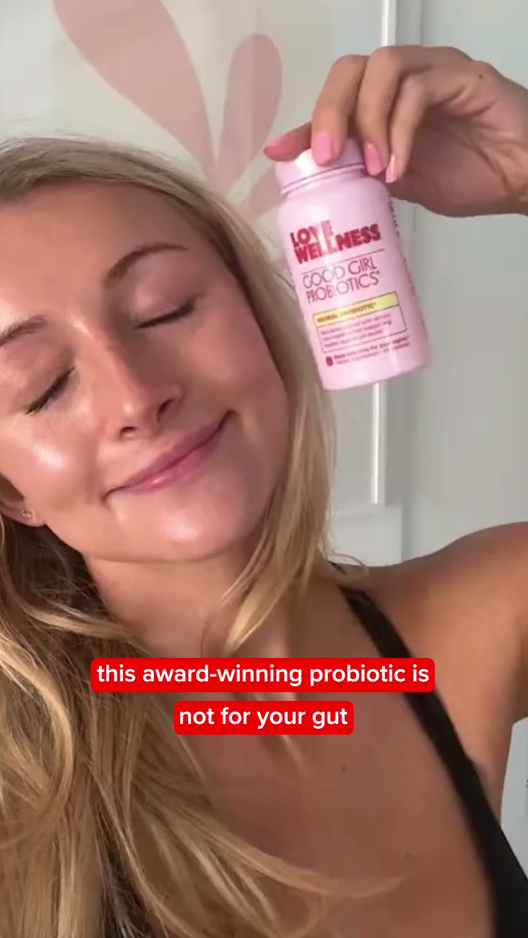 A woman with blonde hair and closed eyes smiles as she holds up a pink bottle of "Love Wellness Good Girl Probiotics." The text overlay on the image reads, "this award-winning probiotic is not for your gut." The woman appears to be a model, and the overall aesthetic suggests a lifestyle or product-focused advertisement.