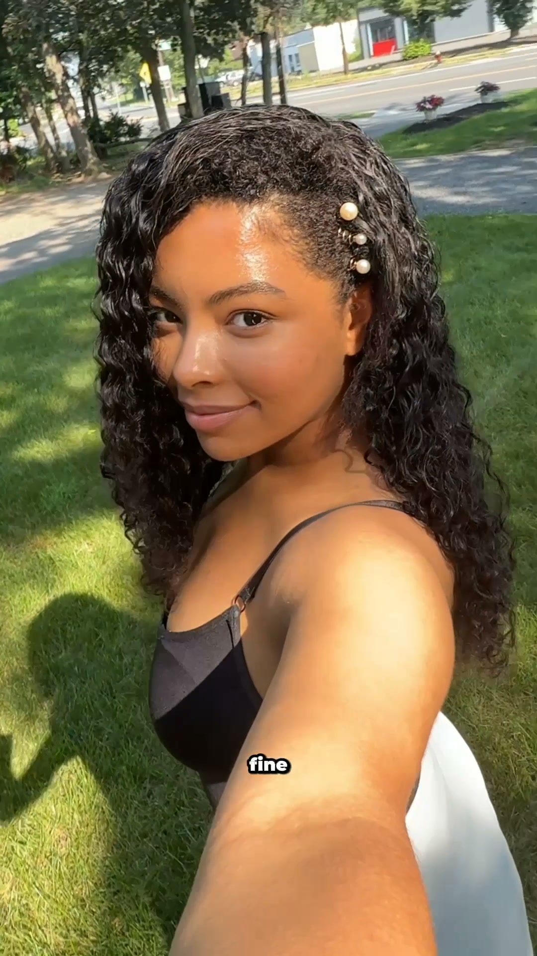 A young woman with curly dark hair is shown from the chest up, looking towards the camera with a slight smile. She is outdoors on a sunny day, with green grass in the foreground and trees and a street in the background. She is wearing a black tank top, and her hair is adorned with pearl hair clips. The word "fine" is overlaid on the image in white text with a black outline.
