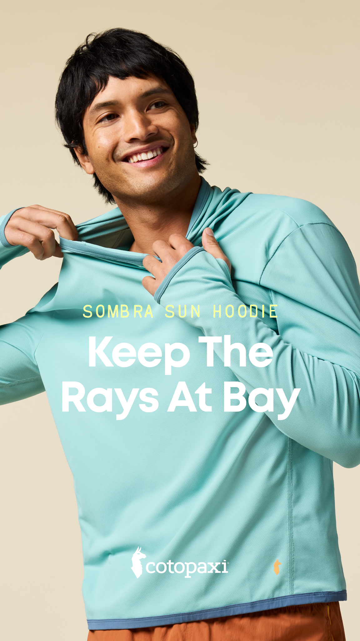 A man with dark hair and a light complexion smiles as he pulls the hood of a light blue long-sleeved shirt over his head. The shirt has "SOMBRA SUN HOODIE" printed in yellow text above the words "Keep The Rays At Bay" in large white font. The Cotopaxi logo is visible on the lower right side of the shirt. The background is a plain, light beige color.