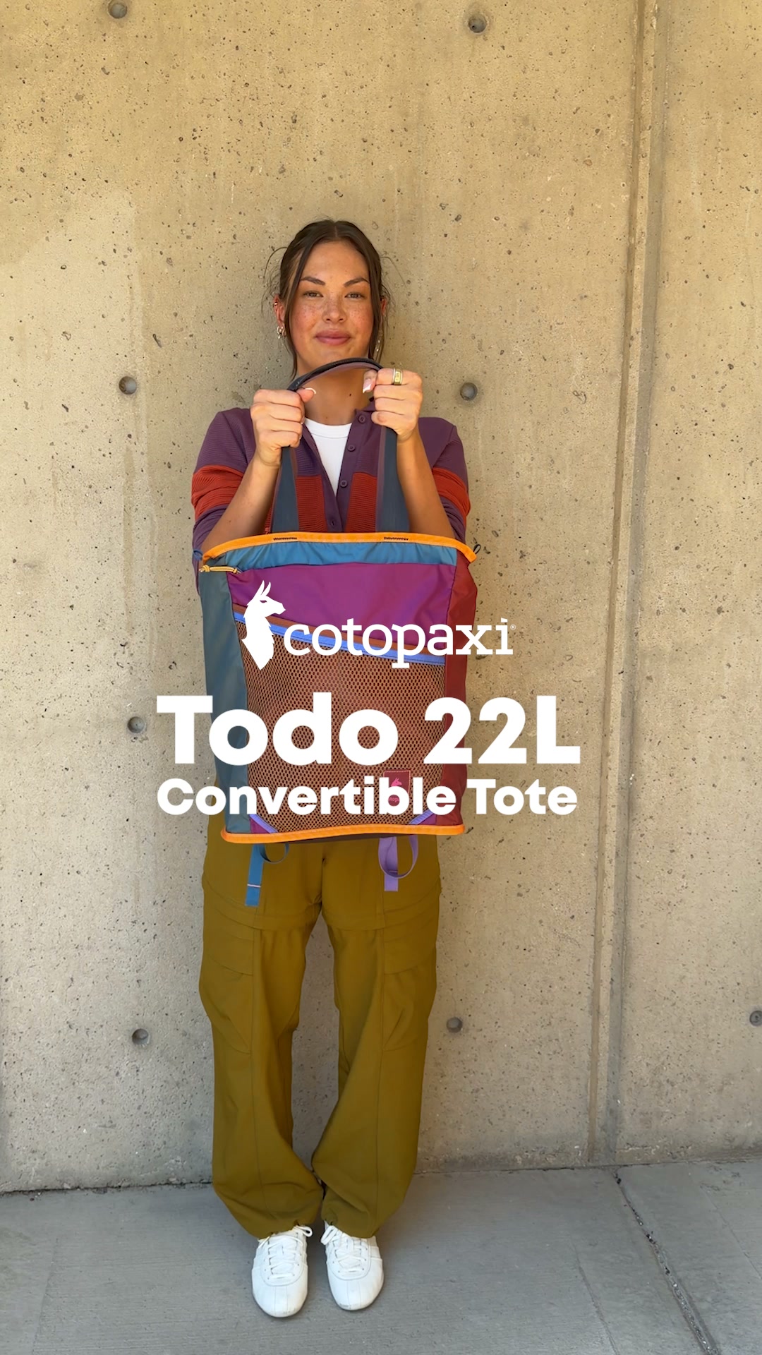 A woman stands against a concrete wall, holding a colorful Cotopaxi convertible tote bag. She is wearing a purple and orange cardigan over a white shirt and olive green cargo pants, with white sneakers. White text overlaid on the image reads "Cotopaxi Todo 22L Convertible Tote." The hook "Never one w/o the other: great day + great gear." is also displayed in white text.