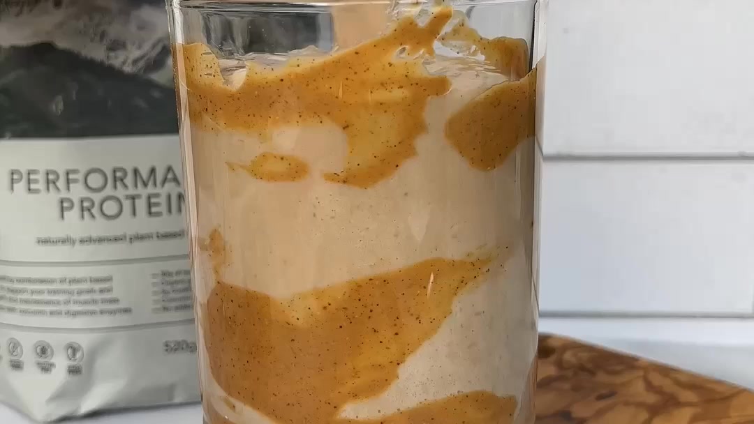 A close-up shot of a glass filled with a creamy, beige-colored smoothie or shake, swirled with a darker, caramel-colored sauce. In the background, a bag of "PERFORMA PROTEIN" is visible, with text indicating it is "naturally advanced plant based". The image focuses on the texture and appearance of the drink, with the protein powder bag serving as context for the product's use.