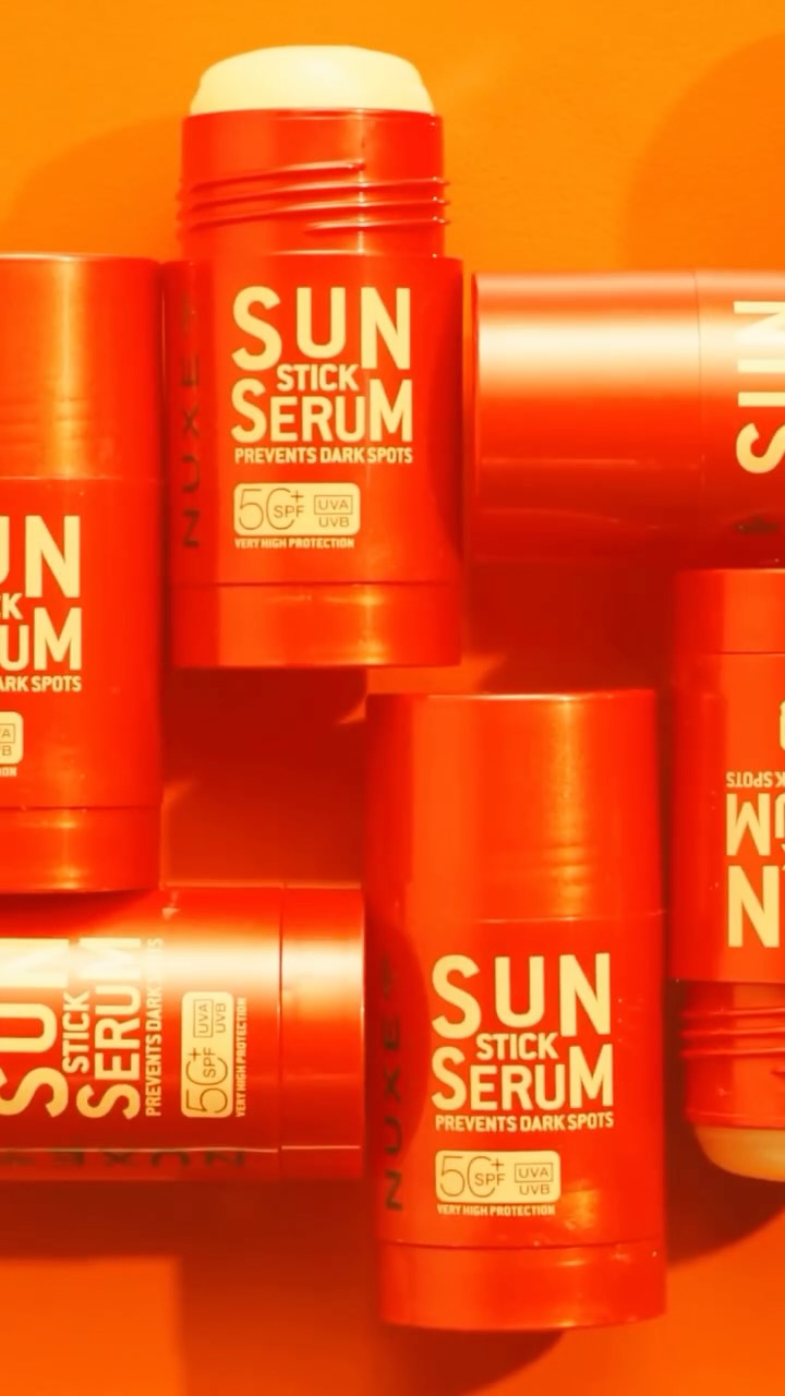 Multiple orange tubes of Nuxe Sun Stick Serum SPF50+ are arranged in a visually appealing, overlapping pattern against an orange background. The text on the tubes reads "SUN STICK SERUM PREVENTS DARK SPOTS" and "VERY HIGH PROTECTION 50+ SPF UVA UVB". The creative highlights the product's dual function as both sun protection and a serum, emphasizing its benefits like anti-aging protection, moisturization, and prevention of dark spots.