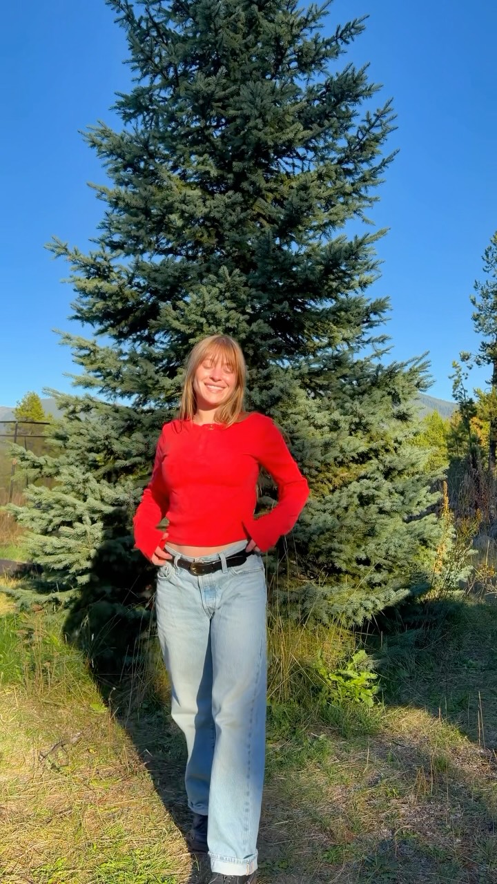 A young woman with blonde hair and bangs, wearing a red long-sleeved shirt and light-wash jeans, stands outdoors in front of a large evergreen tree. She is smiling and looking at the camera, with her hands on her hips. The text "basic tops for layering this fall 🍁" is overlaid at the top of the image. The background shows a clear blue sky and hints of distant mountains and trees. The overall scene suggests a casual, autumnal setting.