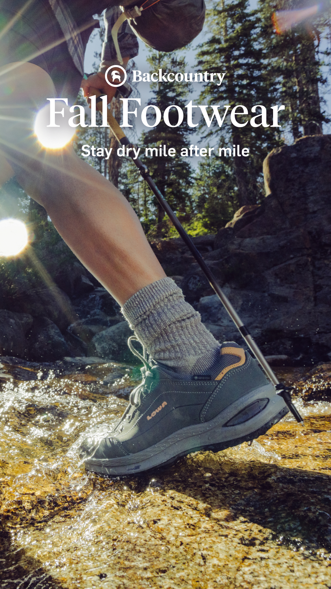 A close-up shot shows a person's foot wearing a hiking shoe and grey sock as they step through shallow, sun-dappled water. The shoe is dark grey with orange accents and the brand name "LOWA" is visible. The text "Backcountry" appears at the top, followed by the headline "Fall Footwear" and the tagline "Stay dry mile after mile." The background features trees and rocks, suggesting an outdoor environment.