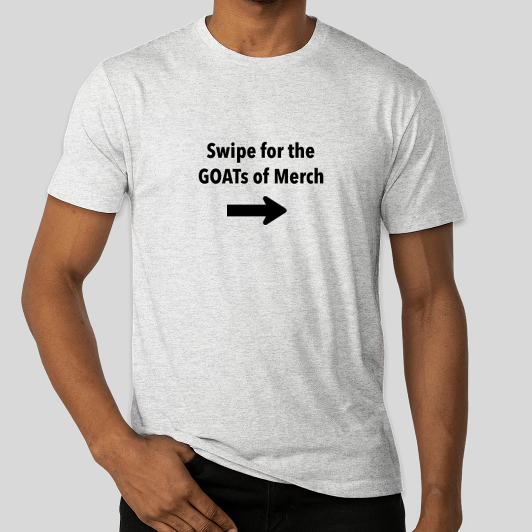 A man wearing a light grey t-shirt is shown from the chest up against a neutral grey background. The t-shirt has black text printed on it that reads "Swipe for the GOATs of Merch" with a black arrow pointing to the right below the text. The man's right hand is casually placed in his front pocket, and his left arm is bent at the elbow, with his hand resting on his chest.