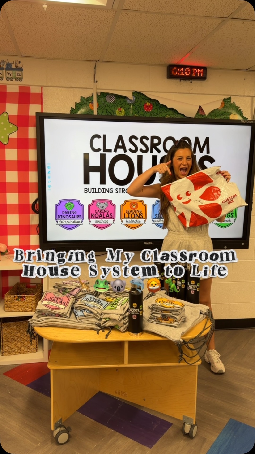 A female teacher stands in a classroom in front of a large screen displaying "CLASSROOM HOUSES". She is holding a white bag with a red logo and pointing at the screen. Below the screen, a wooden cart displays stacks of t-shirts, water bottles, and bags, each representing a different classroom house with animal mascots. The text "Bringing My Classroom House System to Life" is overlaid on the image. The teacher is smiling and appears enthusiastic about her classroom setup.