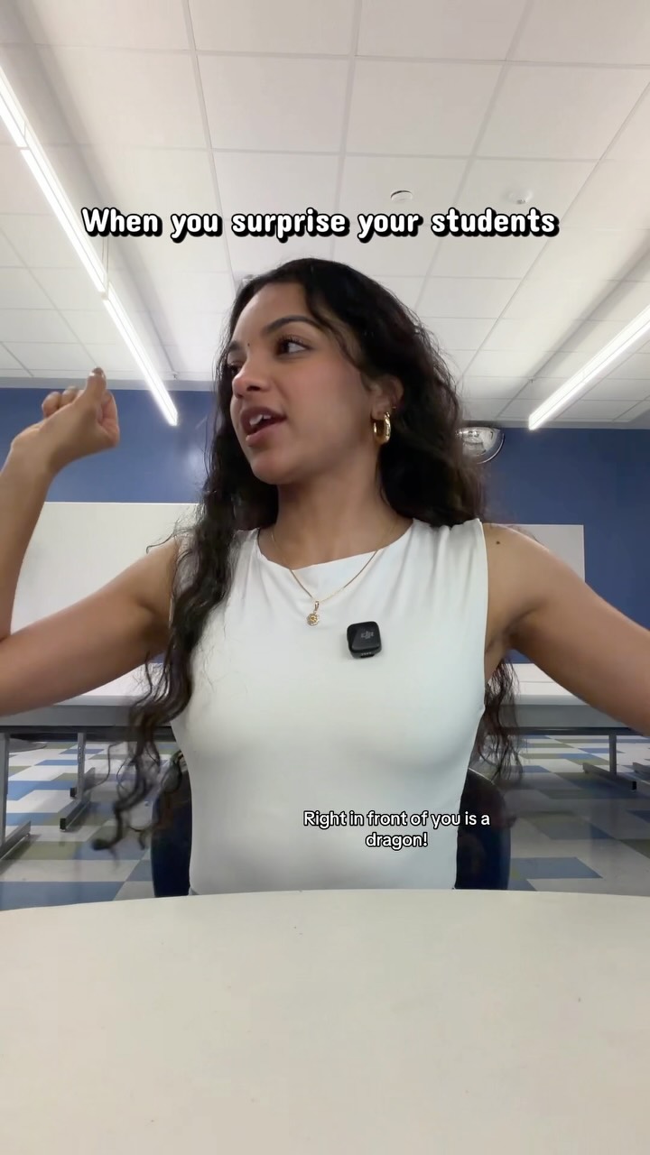 A young woman with dark, curly hair, wearing a white sleeveless top and a gold necklace, is seated at a desk in what appears to be a classroom. Text overlays read "When you surprise your students" at the top and "Right in front of you is a dragon!" near the bottom. She is flexing her bicep and looking off to the side with a playful expression, suggesting a humorous or unexpected scenario.