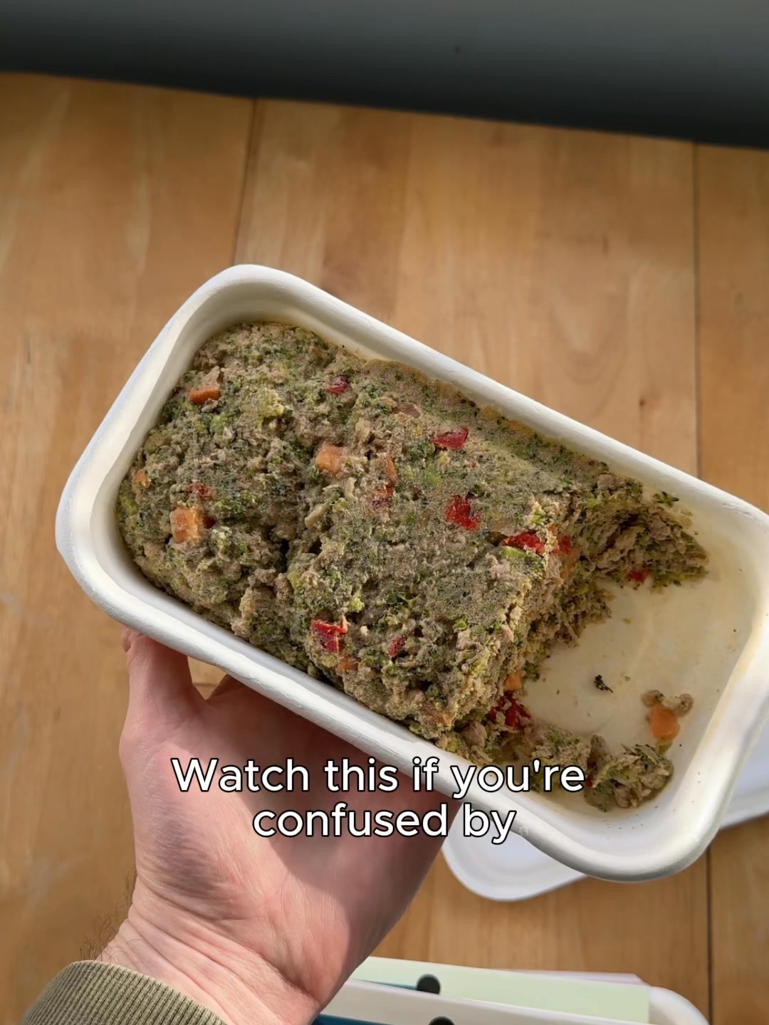 A hand holds a white rectangular container filled with a textured, greenish-brown dog food mixture containing visible pieces of vegetables like broccoli and red peppers. The container is tilted, revealing a portion of the food has been scooped out. White text overlaid on the bottom of the image reads "Watch this if you're confused by". The background is a wooden surface.