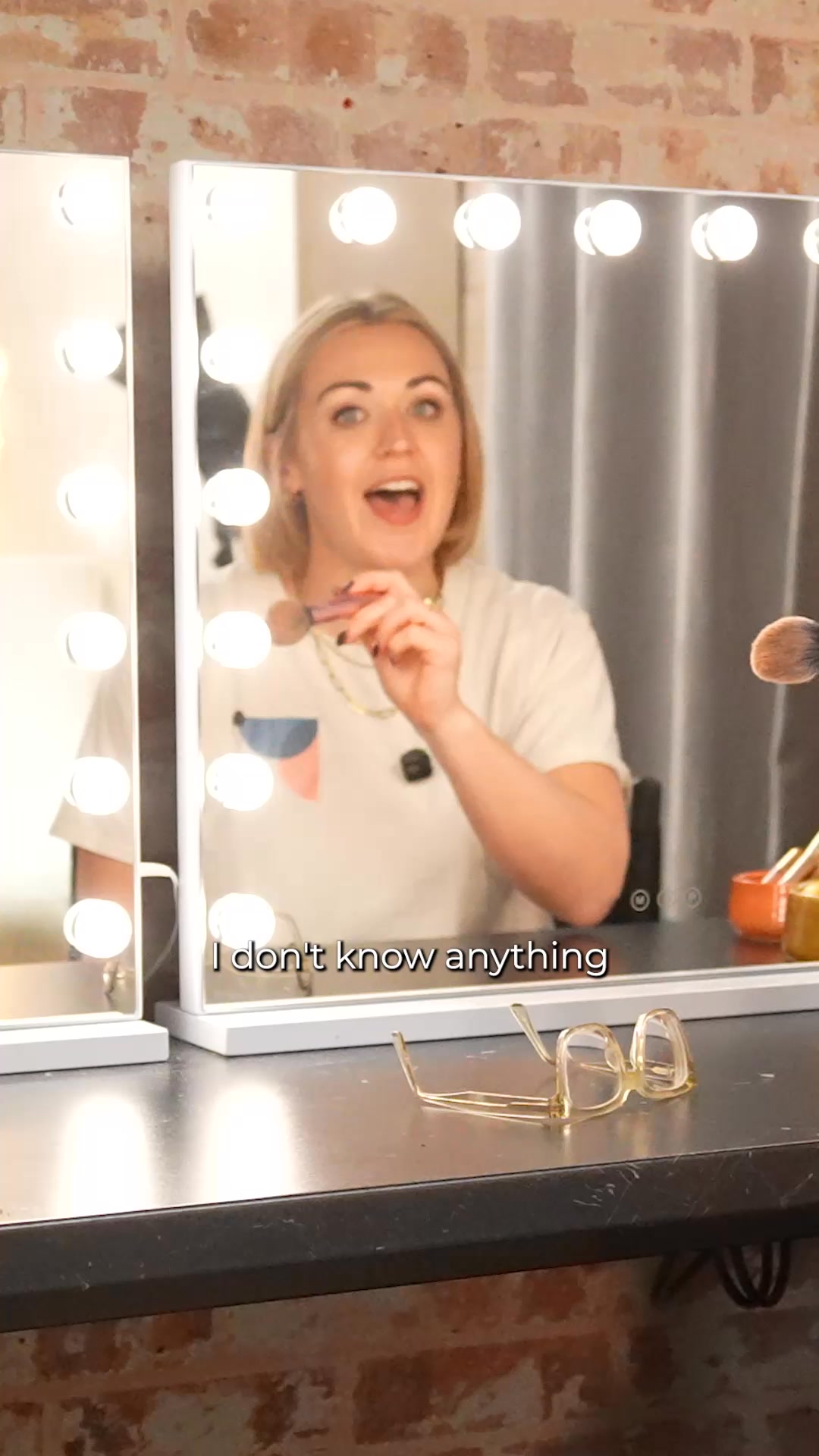 A woman is reflected in a vanity mirror with lights. She is holding a makeup brush and appears to be talking. The text on the screen reads "I don't know anything." In the foreground, a pair of glasses rests on the counter. The overall scene suggests a moment of self-reflection or perhaps a humorous admission about not knowing something, possibly related to makeup or a personal transformation.