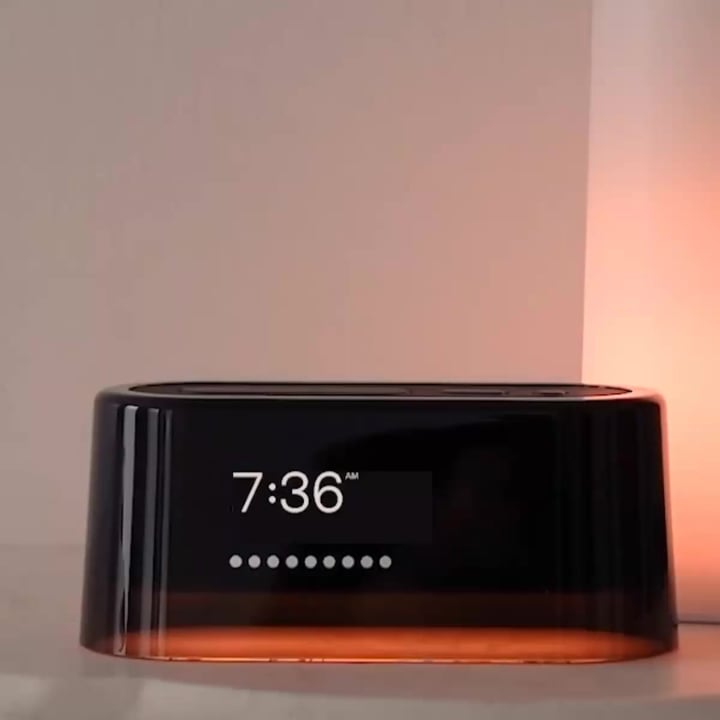A close-up shot of a black Loftie alarm clock displaying the time "7:36 AM" with a row of white dots below it. The clock has an orange glowing base. The text "Get your best sleep yet" is overlaid on the image, followed by a sentence about the product's benefits and a phone emoji.