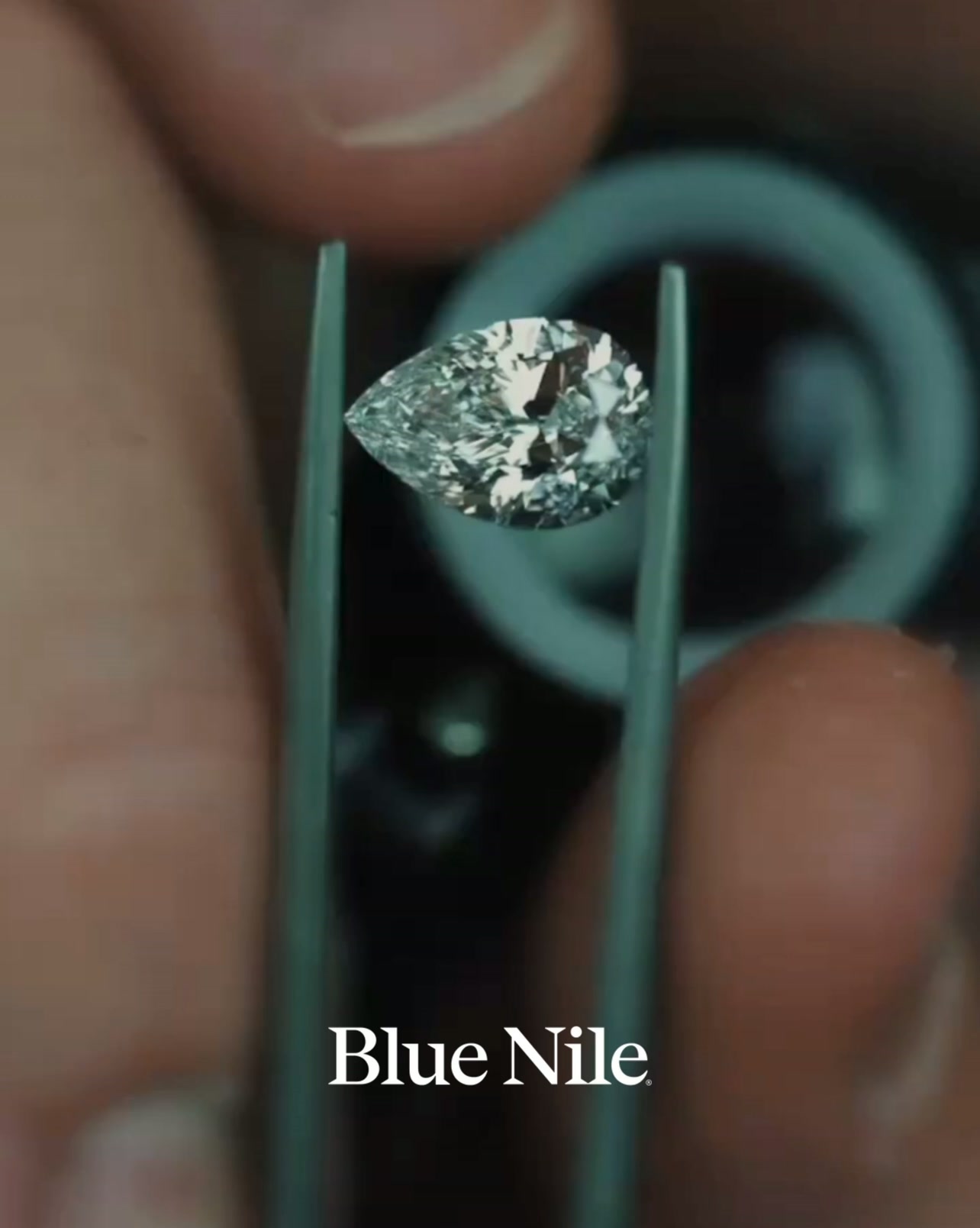 A close-up shot shows a pear-shaped diamond being held by tweezers. The diamond is brilliant and reflects light. Below the diamond, the Blue Nile logo is displayed in white text. The background is blurred and dark, with hints of blue and grey. The text overlay introduces the concept of six diamond shapes and their legacy.