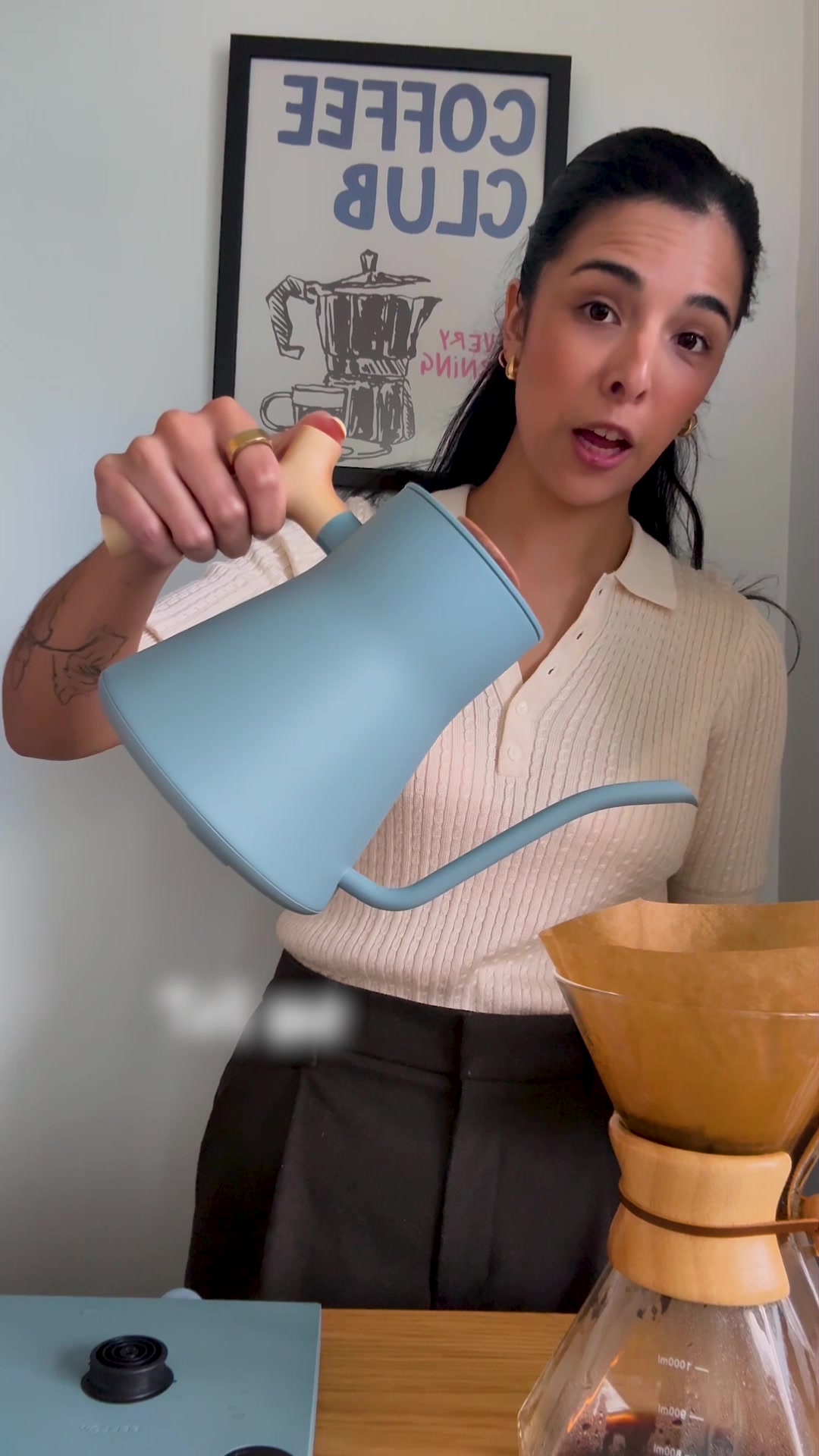 A woman with dark hair, wearing a cream-colored polo shirt, is holding a light blue gooseneck kettle over a pour-over coffee maker. She is in the process of pouring water into the coffee filter. Behind her, a framed poster with the words "COFFEE CLUB" in blue lettering is visible. The text overlay on the image explains the features of the Stagg EKG kettle, highlighting its ability to maintain precise water temperatures and time brews, while also noting its aesthetic appeal.
