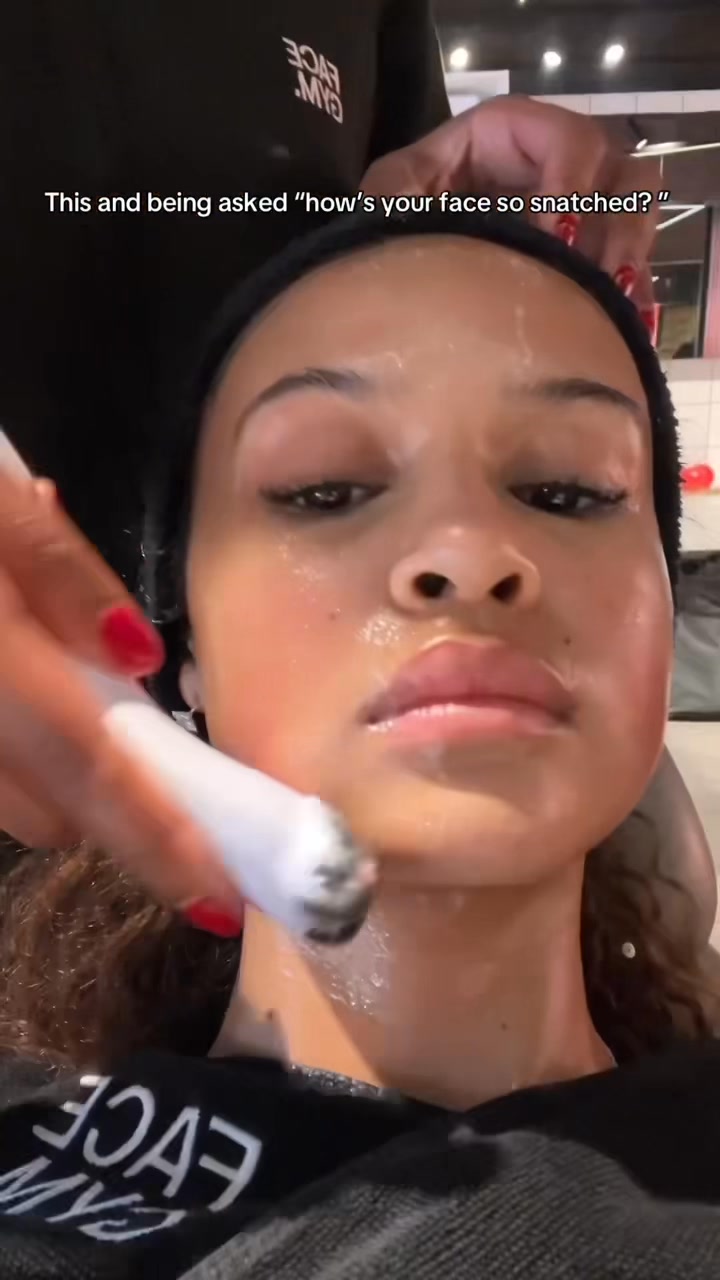 A close-up shot of a young woman's face as she receives a facial treatment. Her eyes are closed, and she has a serene expression. A hand with red nail polish is holding a tool near her jawline, applying a white substance. Text overlayed on the image reads, "This and being asked 'how's your face so snatched?'" The background shows a blurred gym-like environment with the logo "FACEGYM" visible on a black t-shirt.