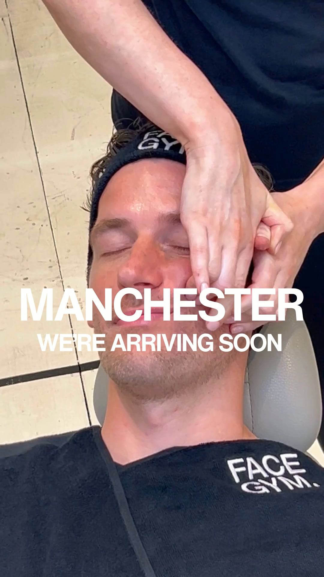 A close-up shot of a man with his eyes closed, receiving a facial massage. Large white text overlays the image, reading "MANCHESTER WE'RE ARRIVING SOON". The man is wearing a black headband with "GYM" visible and a black t-shirt with "FACE GYM." printed on it. The overall impression is an announcement of a new location opening.