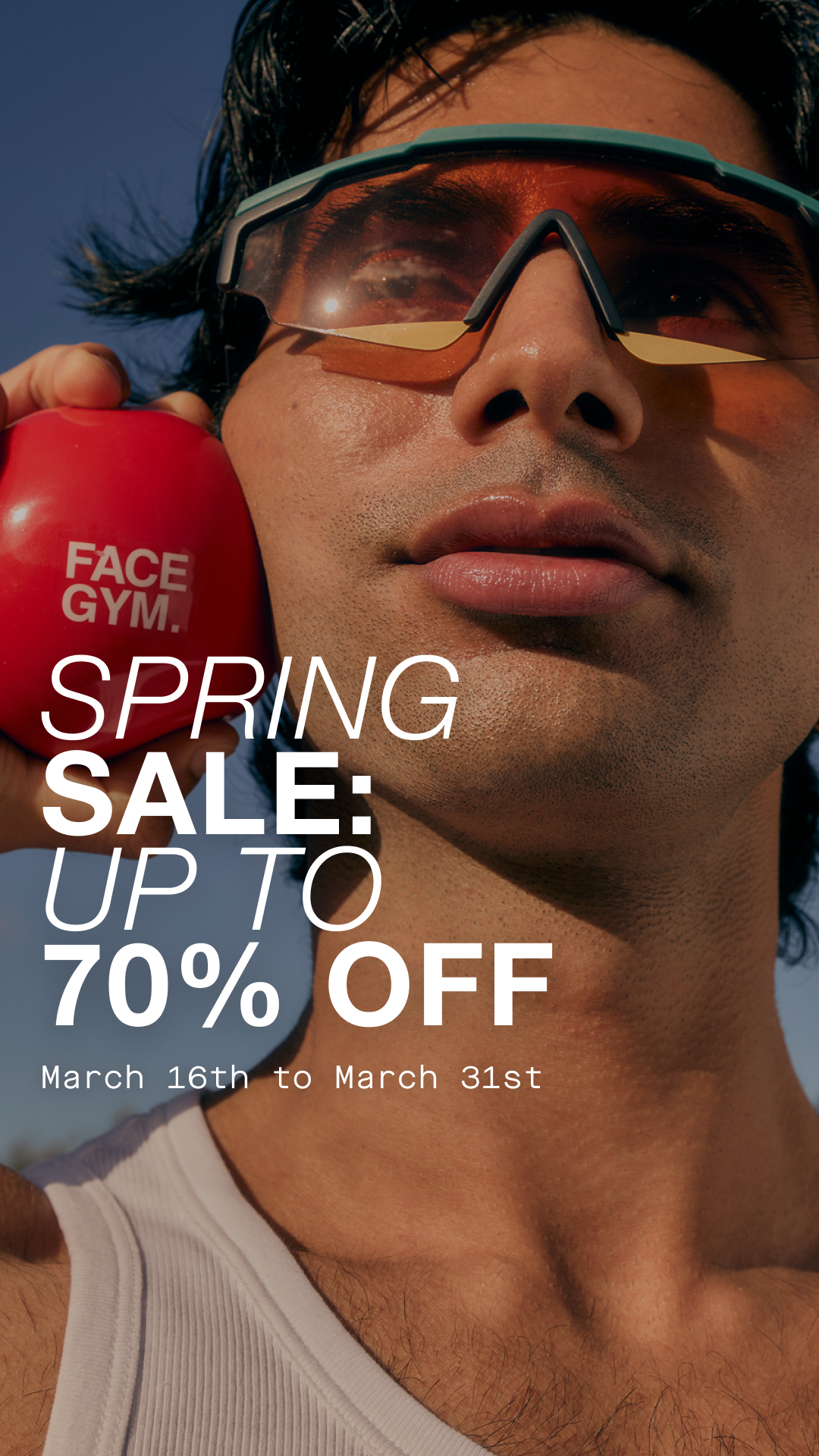 A close-up, low-angle shot of a man wearing sunglasses and a white tank top, holding a red FaceGym ball to his cheek. Large white text overlays the image, announcing "SPRING SALE: UP TO 70% OFF" and the dates "March 16th to March 31st". The man's face is partially obscured by the sunglasses and the angle, with the sun glinting off the lenses. The overall impression is a stylish, promotional image for a sale event.