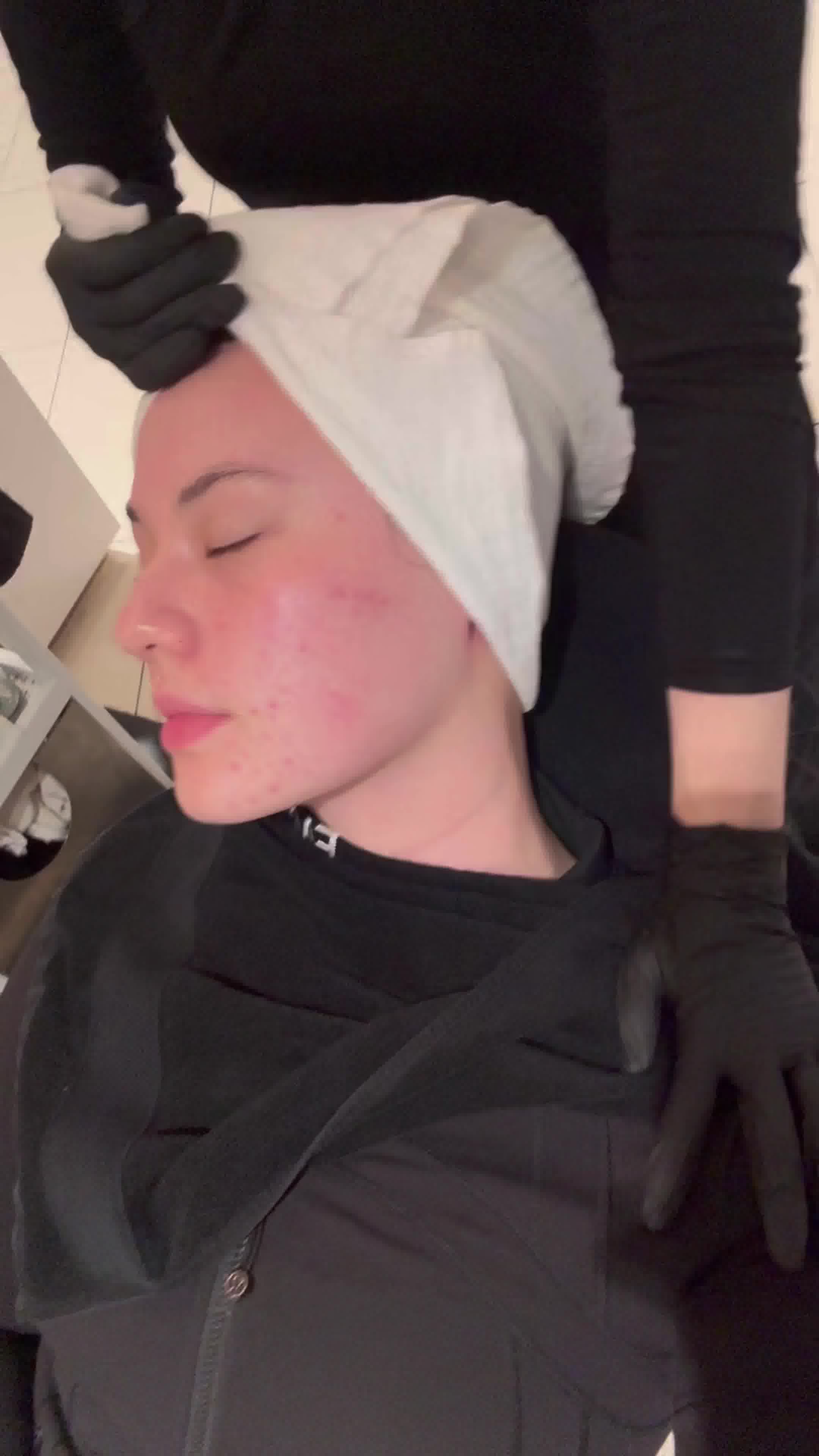 A close-up profile shot of a young woman with her eyes closed, receiving a facial treatment. Her face has some redness and blemishes. A person wearing black gloves is gently placing a white towel on her head. The text overlay reads: "Your FACEGYM Workout just got an upgrade 😌 Supercharged formulas. Instant results." The overall impression is one of pampering and advanced skincare.