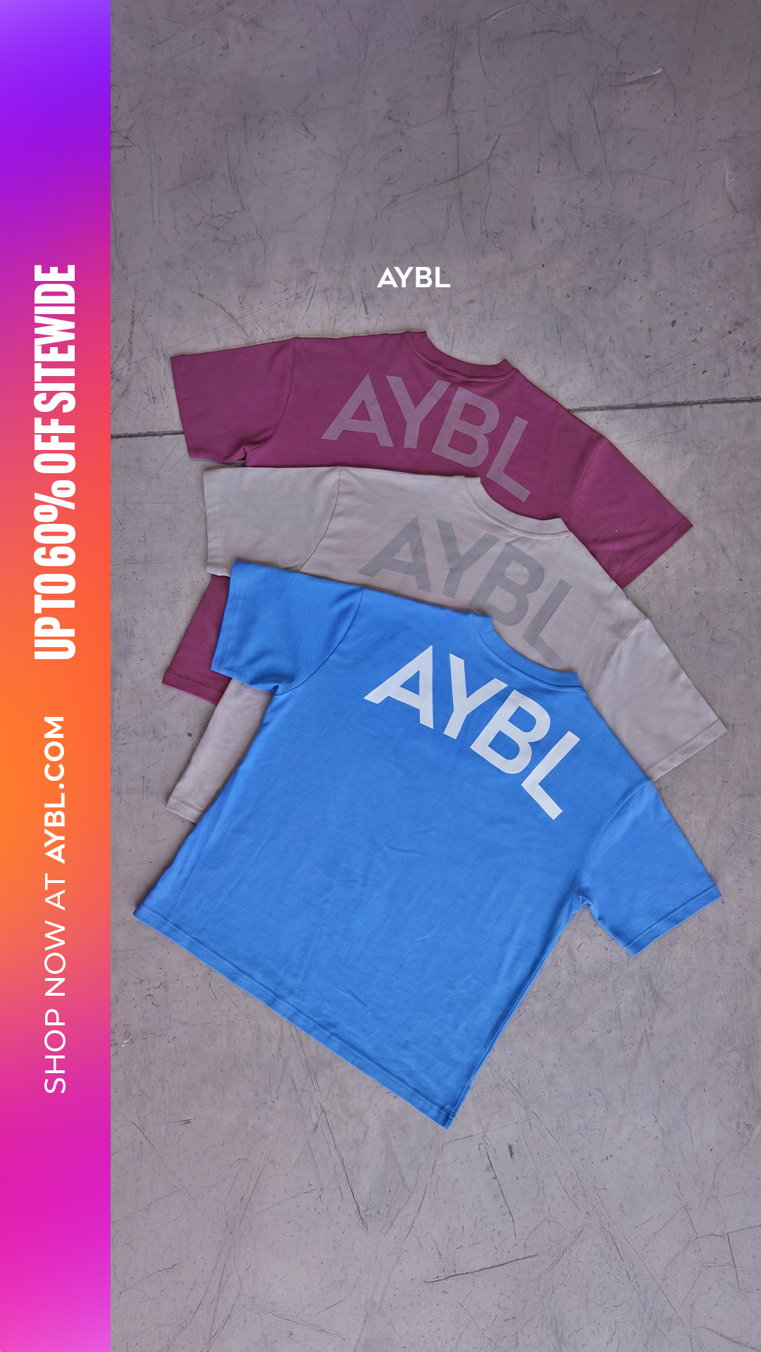 Three t-shirts are stacked on top of each other on a grey, textured surface. The t-shirts are in shades of purple, beige, and blue, with the brand logo 'AYBL' printed in a lighter shade on the purple and beige shirts, and in white on the blue shirt. A vertical gradient bar on the left side of the image contains white text that reads 'SHOP NOW AT AYBL.COM' and 'UP TO 60% OFF SITEWIDE'.