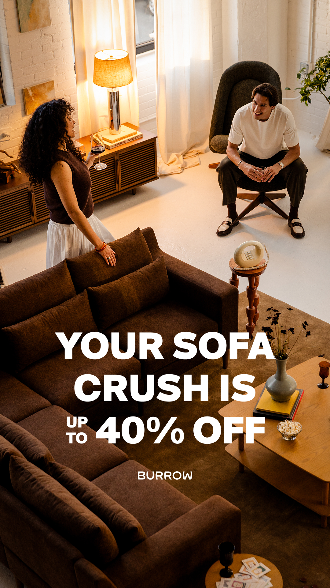 A man and a woman are in a modern living room. The woman is standing near a large brown sectional sofa, holding a glass of wine. The man is sitting in a chair, looking away from the camera and smiling. Large white text overlays the image, reading "YOUR SOFA CRUSH IS UP TO 40% OFF" and "BURROW" at the bottom. The scene is well-lit with natural light coming from large windows.
