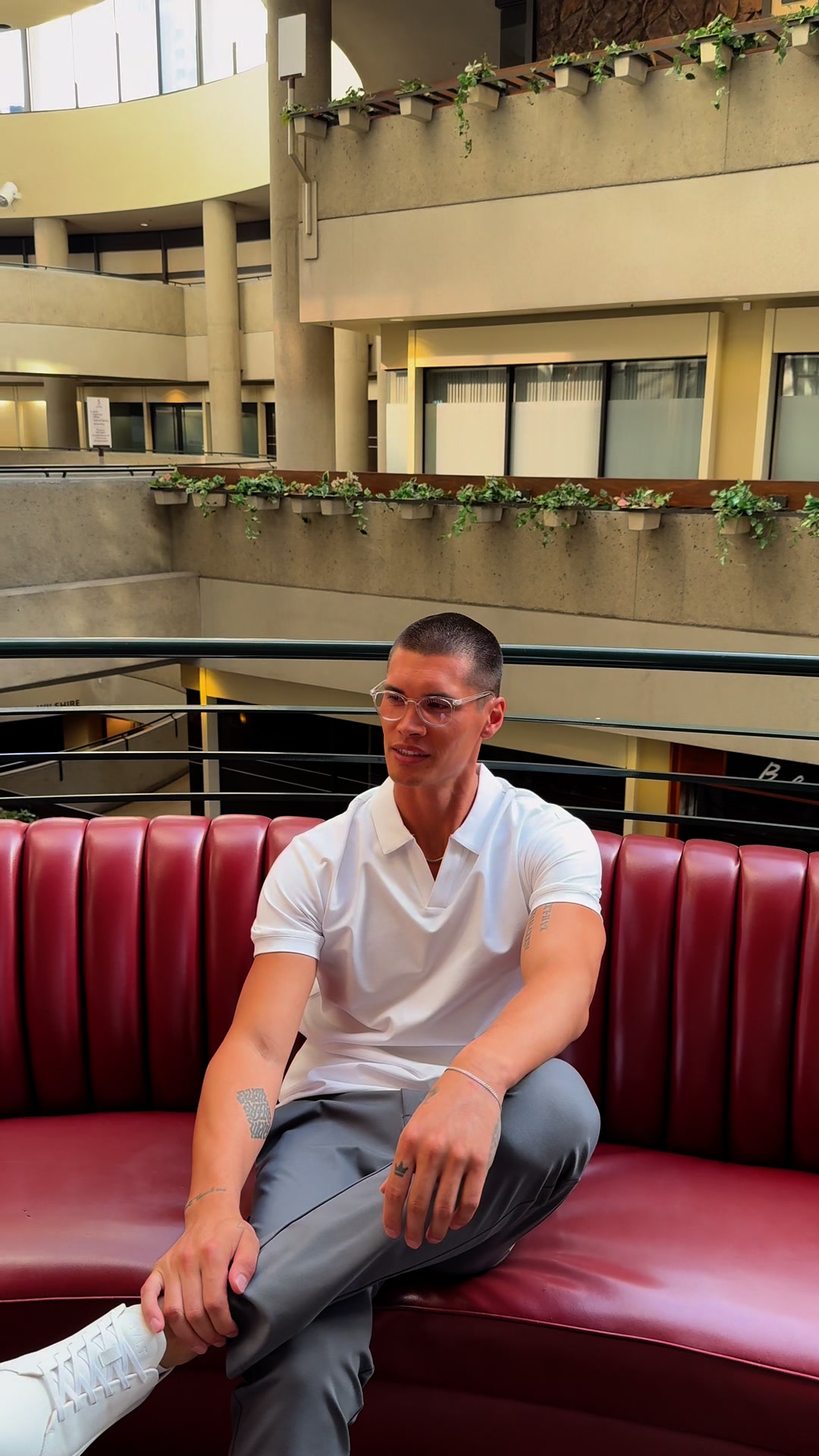 A man with short hair and glasses sits on a red leather couch in a modern atrium. He is wearing a white polo shirt and grey pants. Text overlays the image, listing benefits such as "Over 100K+ 5-Star Reviews" and "Loved by Forbes, Entrepreneur, Men's Journal." The ad promotes premium basics and highlights various offers like free gifts and free shipping.