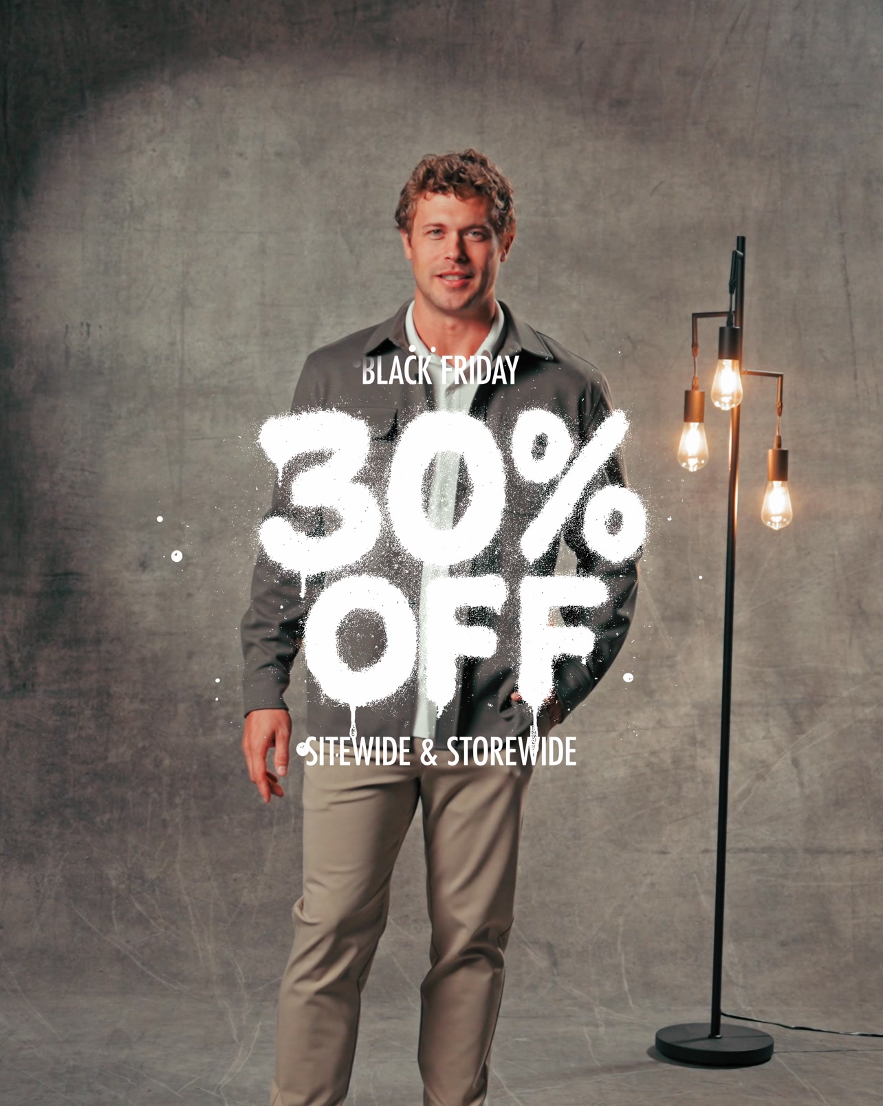A man stands against a textured grey backdrop, wearing a grey jacket over a light-colored shirt and tan pants. Large, white, graffiti-style text is spray-painted across the image, reading "30% OFF" with "SITIDEWIDE & STOREWIDE" below it. The text "BLACK FRIDAY" is also visible above the "30% OFF" graphic. A floor lamp with three exposed bulbs stands to the right of the man.