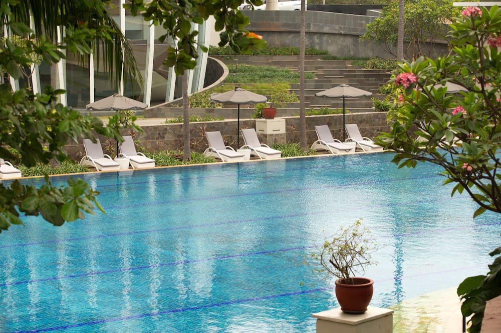 A serene swimming pool area with lounge chairs and umbrellas is shown. The water is a vibrant blue with darker blue lines. Lush green trees and foliage frame the scene, with a modern building visible in the background. On-screen text asks, "Du möchtest flexibel sein?" (Do you want to be flexible?) and mentions that many hotels can be canceled for free on the Expedia app.