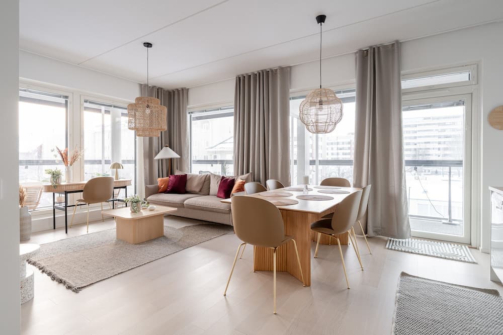 A bright, modern living and dining area is shown. The space features a comfortable sofa, a dining table with chairs, and large windows with grey curtains. Text overlays the image, stating "Encuentra ofertas de hotel para tu próximo viaje con Expedia. Busca opciones flexibles con cancelación gratis." The overall impression is one of a stylish and inviting home.