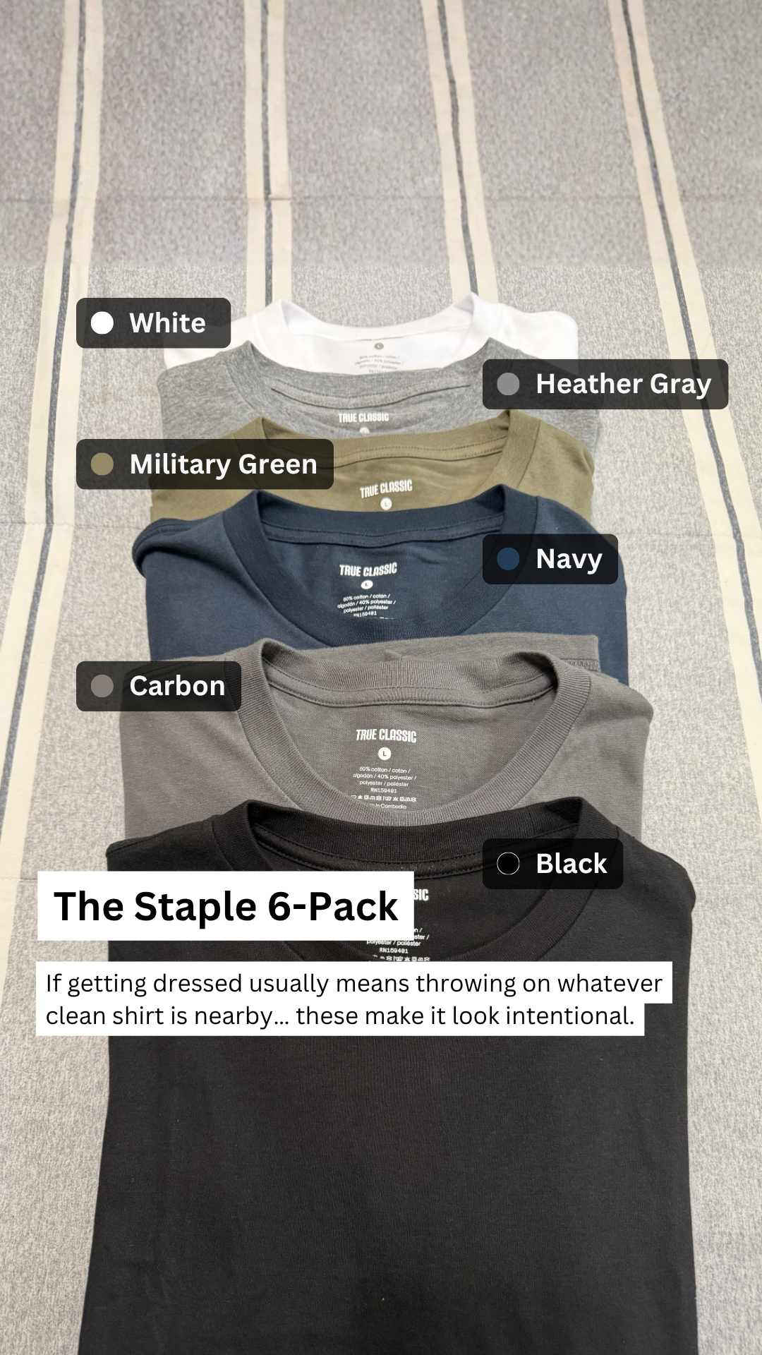 A stack of five True Classic t-shirts in different colors is shown from the neckline down. The colors are White, Heather Gray, Military Green, Navy, and Carbon. A sixth black t-shirt is laid out flat at the bottom of the stack. Text overlays identify the colors of the shirts. A larger text box at the bottom reads "The Staple 6-Pack" and below that, "If getting dressed usually means throwing on whatever clean shirt is nearby... these make it look intentional." The background is a striped fabric.