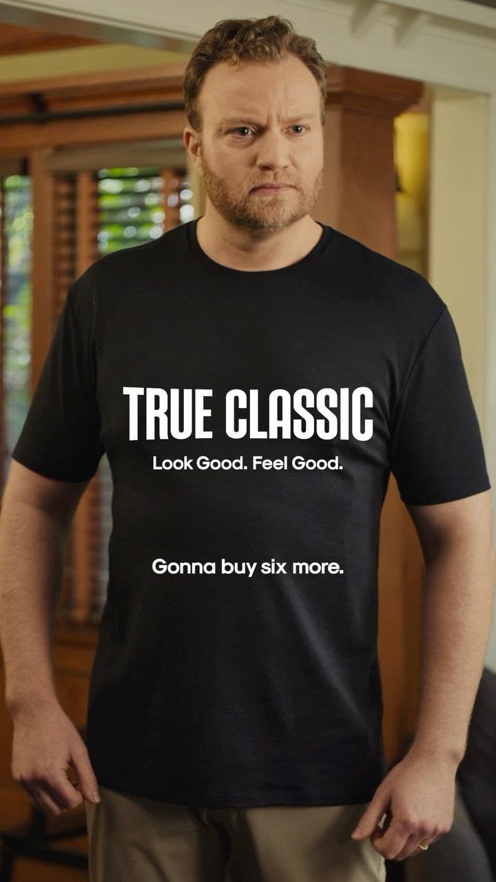 A man wearing a black t-shirt stands facing the camera. The t-shirt has white text that reads "TRUE CLASSIC" in large font, followed by "Look Good. Feel Good." and then "Gonna buy six more." The man has a slightly skeptical or unimpressed expression. The background appears to be an indoor setting with wooden blinds and furniture, suggesting a home environment. The overall impression is that the man is impressed with the t-shirt and intends to purchase more.