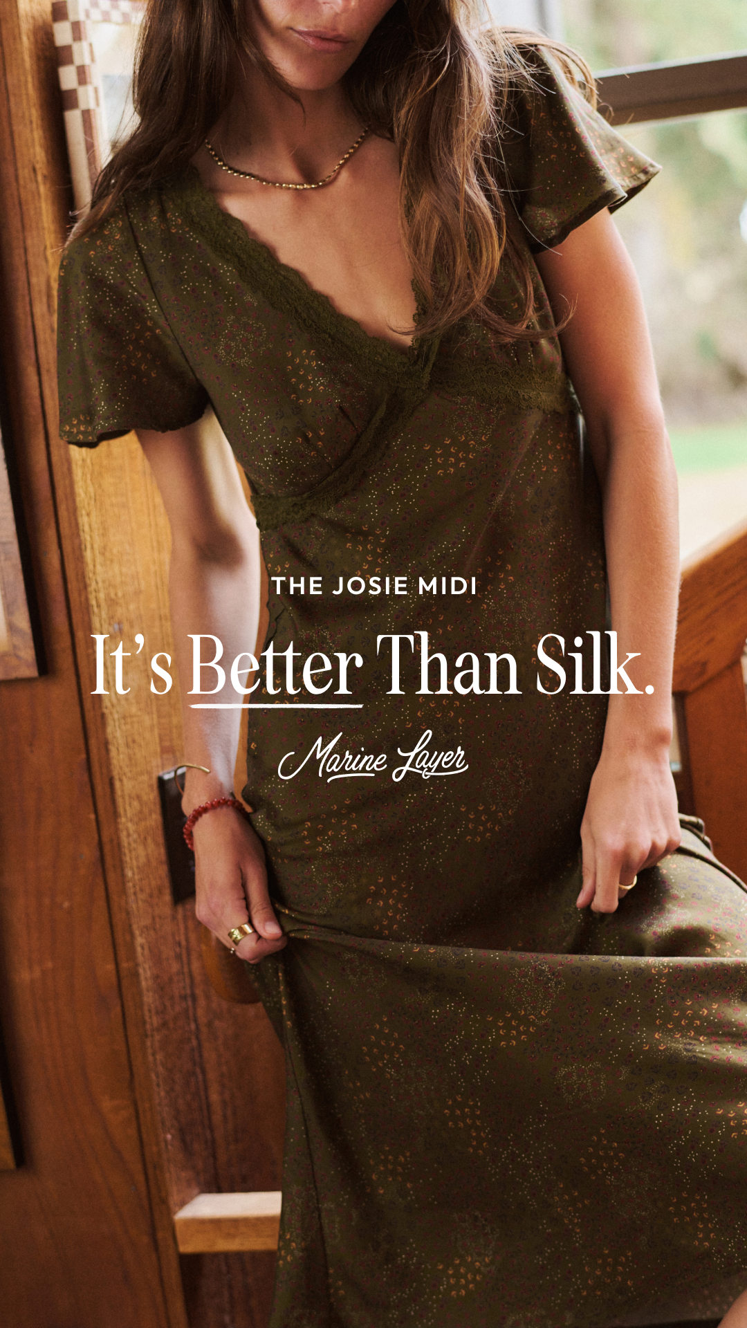 A woman wearing a green, patterned midi dress with lace trim is leaning against a wooden structure near a window. The text "THE JOSIE MIDI" appears above the main headline, "It's Better Than Silk." Below the headline, the brand name "Marine Layer" is written in a script font. The image focuses on the texture and drape of the dress, suggesting comfort and quality.