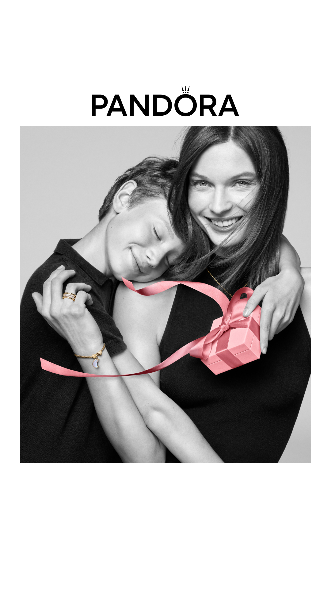 A black and white image shows two women embracing. The woman on the right is smiling broadly at the camera, her arm around the woman on the left. The woman on the left has her eyes closed and her head resting on the other woman's shoulder. A pink ribbon is draped around them, and a small pink gift box is held by the woman on the right. The Pandora logo is displayed prominently at the top of the image. The text "Wenn sie dein Ein und Alles ist – schenk ihr den Mond. #BeLove" is overlaid on the image.