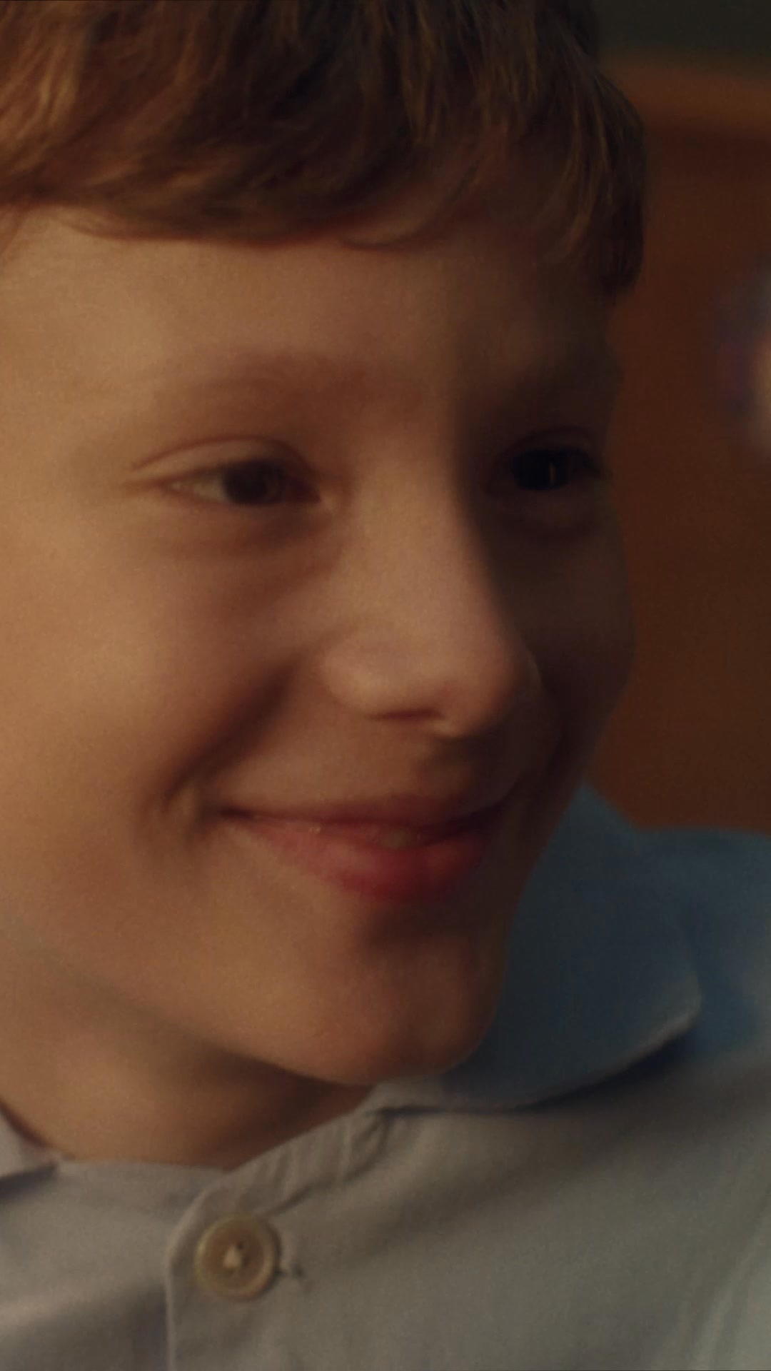 A close-up shot focuses on the face of a young boy with reddish-brown hair, who is smiling warmly. He is wearing a light blue collared shirt with a button at the neck. The background is softly blurred, suggesting an indoor setting. Overlaid on the image is text that reads "Verschenke etwas, das für immer in Erinnerung bleibt." followed by the hashtag "#BeLove", encouraging the gifting of a memorable item.
