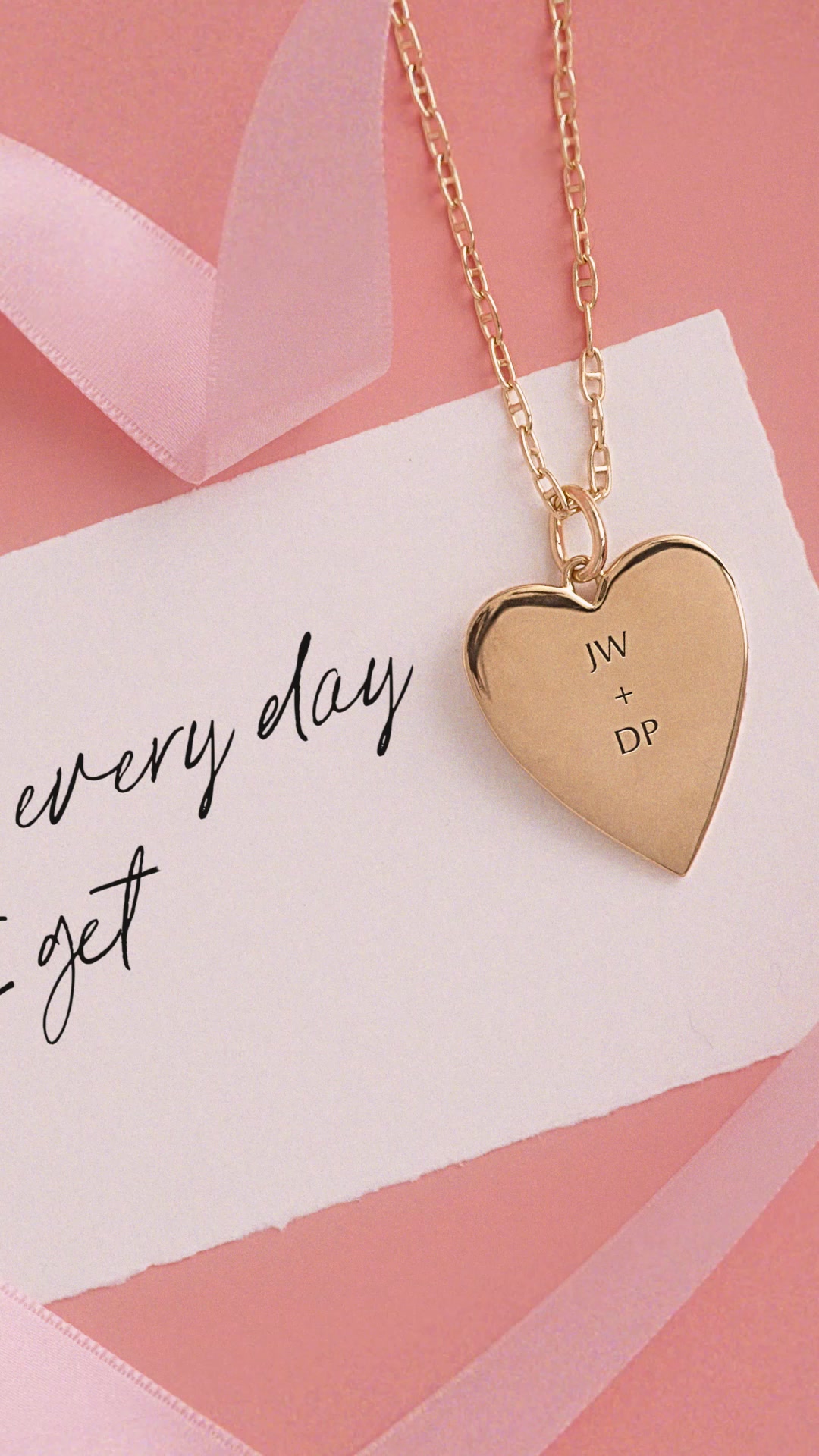 A close-up shot of a gold heart-shaped pendant necklace with the initials "JW + DP" engraved on it. The necklace is resting on a white card with handwritten text that reads "every day I get". A pink ribbon is visible in the background, adding a soft, romantic touch. The overall aesthetic is elegant and sentimental, suggesting a gift for a special occasion.