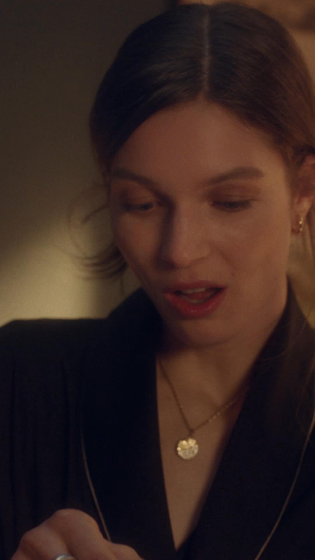 A close-up shot of a young woman with light brown hair, wearing a black robe and a gold pendant necklace. She is looking down with a soft smile and her mouth slightly open. The background is dimly lit and out of focus, creating an intimate atmosphere. The text "Give a gift they’ll always remember. #BeLove" is overlaid on the image.