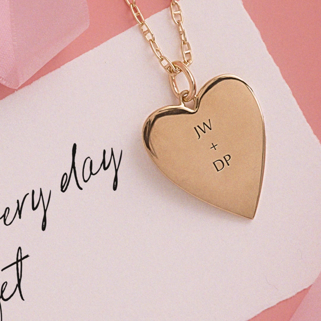 A close-up shot focuses on a gold heart-shaped locket necklace resting on a white card with handwritten text. The locket is engraved with the initials "JW + DP". The background is a soft pink, with a hint of pink ribbon on the left. The handwritten text on the card reads "ery day" and "et". The overall aesthetic is romantic and sentimental, suggesting a personalized gift for a special occasion.