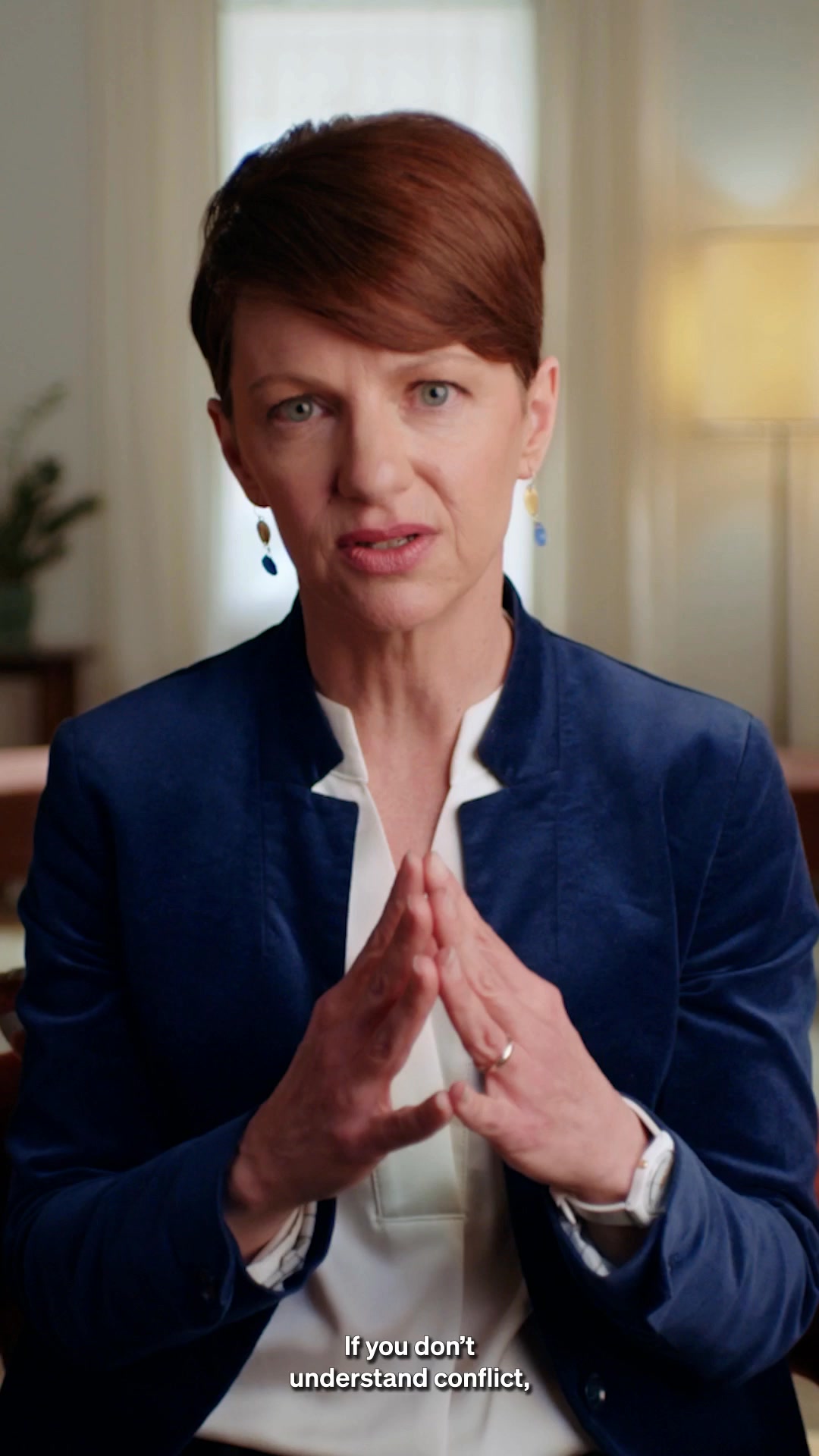 A woman with short, reddish-brown hair and blue eyes, wearing a dark blue velvet blazer over a white blouse, is shown from the chest up. She is looking directly at the camera with her hands clasped in front of her. White text overlays the bottom of the image, reading "If you don't understand conflict,". The background is softly blurred, suggesting an indoor setting with a lamp and curtains. The woman appears to be a founder or expert speaking directly to the viewer.