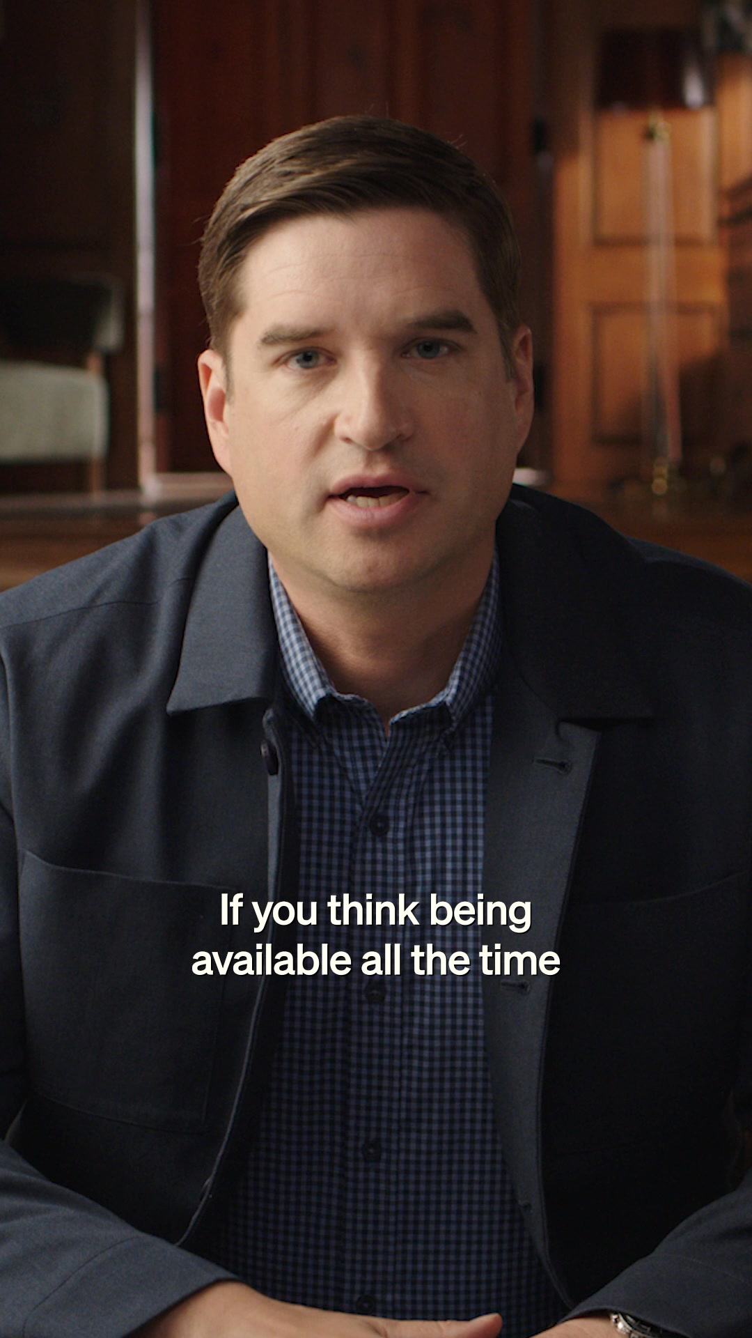 A man in a dark blue jacket over a blue checkered shirt is looking directly at the camera. White text is overlaid on the bottom half of the image, reading "If you think being available all the time". The background appears to be an indoor setting with wooden furniture and paneling.