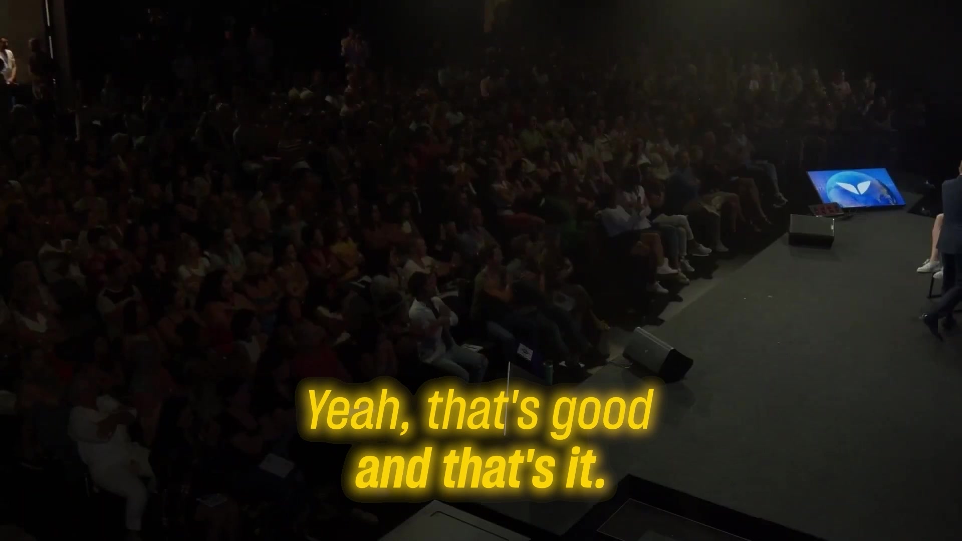 A wide shot shows a large audience seated in a dark auditorium, facing a stage. On the stage, a screen displays a blue logo with white wings. Yellow text with a neon glow is overlaid on the bottom center of the image, reading "Yeah, that's good and that's it." The overall impression is of a presentation or event where a speaker has just concluded a point.