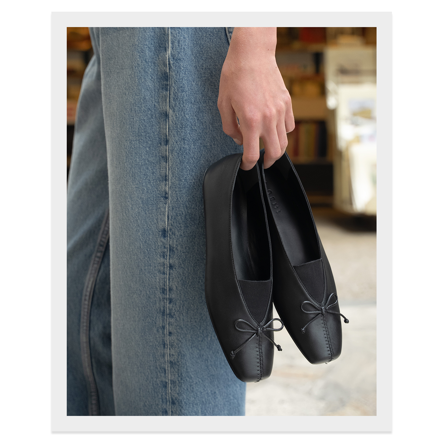 A close-up shot shows a person holding a pair of black ballet flats. The shoes have a square toe and a small bow detail on the vamp. The background is blurred, showing bookshelves and a hint of a store interior. The on-screen text explains that these flats are a new take on a favorite style, featuring a bow instead of straps.