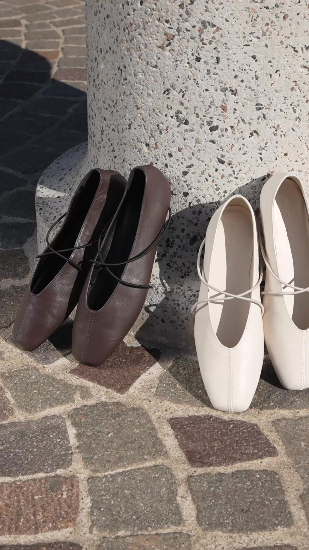 Two pairs of ballet flats are displayed on a textured stone surface. The pair on the left is a deep brown, and the pair on the right is a creamy off-white. Both pairs feature thin, elastic straps that cross over the top of the foot. The text "Our butter-soft flat is our #1 seller for a reason." is overlaid on the image.