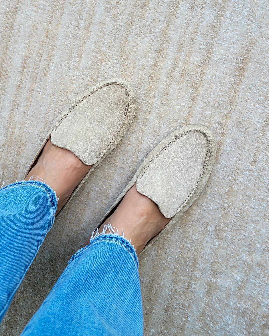 A top-down shot shows a person's feet wearing light beige suede loafers and blue jeans with frayed hems. The loafers are positioned on a textured, cream-colored carpet. The text overlay reads, "The Felize Mule feels like a slipper but looks so much more polished. It's the perfect work-from-home (or anywhere) shoe."