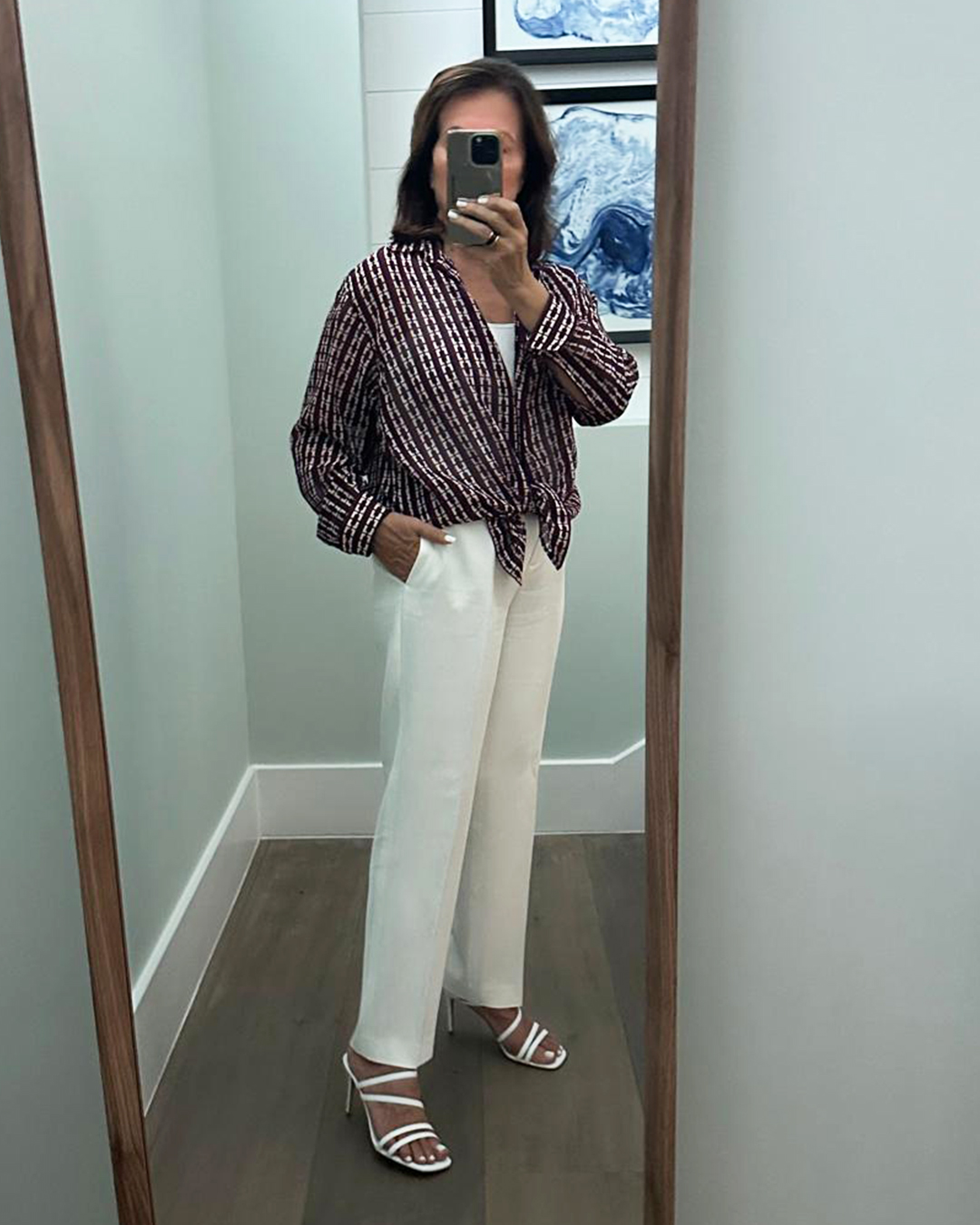 A woman stands in front of a mirror, taking a selfie in a fitting room. She is wearing a striped tie-front blouse and white wide-leg pants with white strappy heels. The text overlay shares a personal anecdote about ruining a pair of shoes on a bike ride and expresses happiness that they are back in stock for the summer.