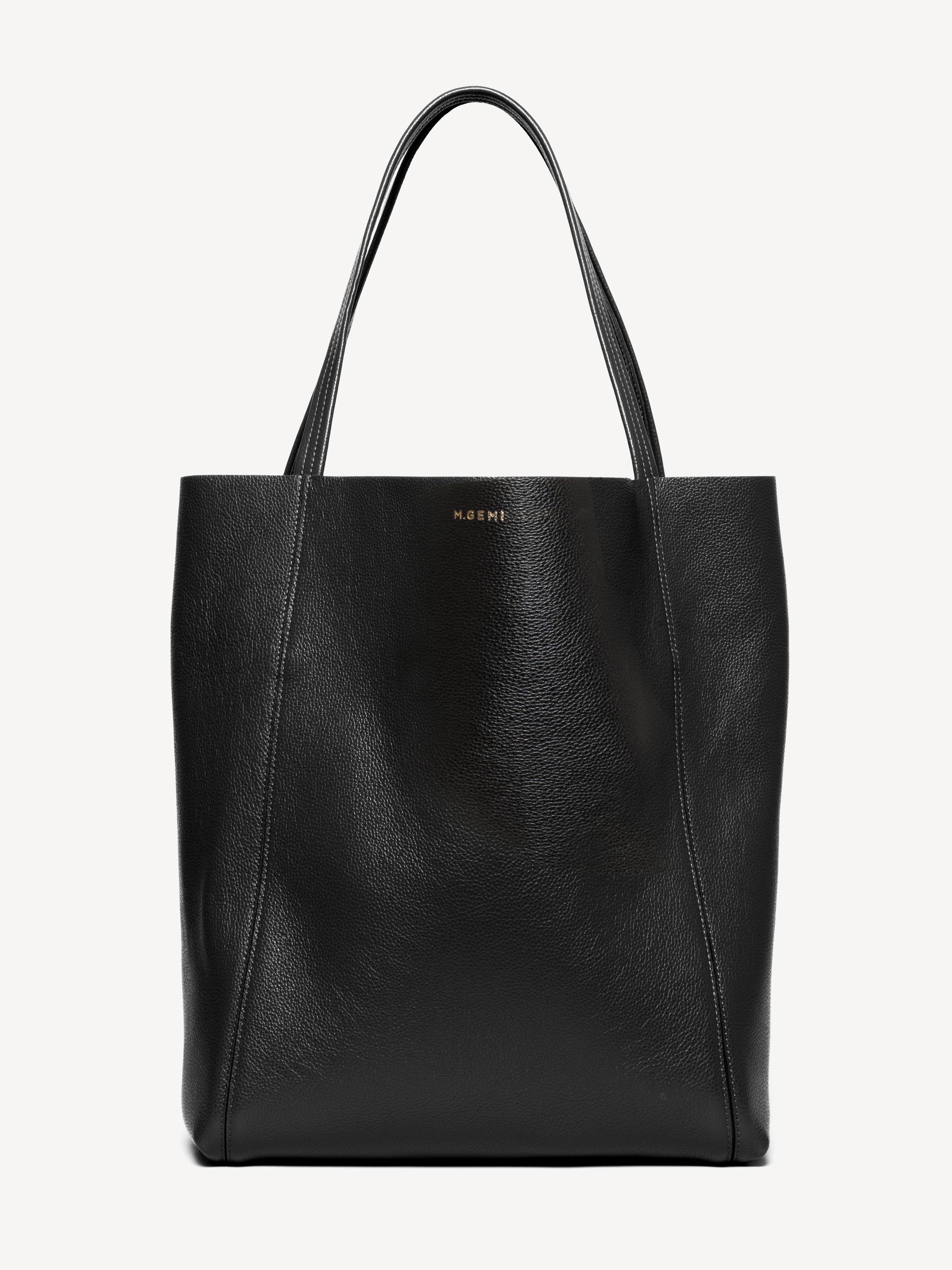 A clean product shot of a black leather tote bag, the M.Gemi Lola, is presented against a plain, light grey background. The bag is shown from the front, with its handles gracefully curved upwards. Subtle white stitching details are visible along the seams of the bag. The brand name 'M.GEMI' is embossed in gold lettering on the front of the bag. The on-screen text, placed at the top of the image, highlights the bag's Italian leather craftsmanship and its versatile, convertible sizing.