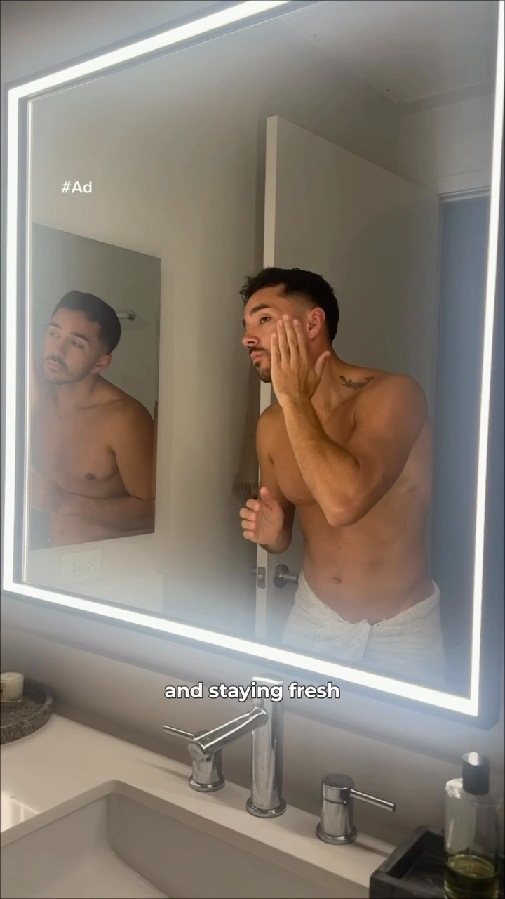 A man is shown in a bathroom, looking into a brightly lit mirror and applying lotion to his face. He is shirtless and wearing a towel around his waist. The reflection in the mirror shows him from the chest up. Text overlay on the bottom of the screen reads "and staying fresh". The hashtag "#Ad" is visible in the top left corner of the mirror.