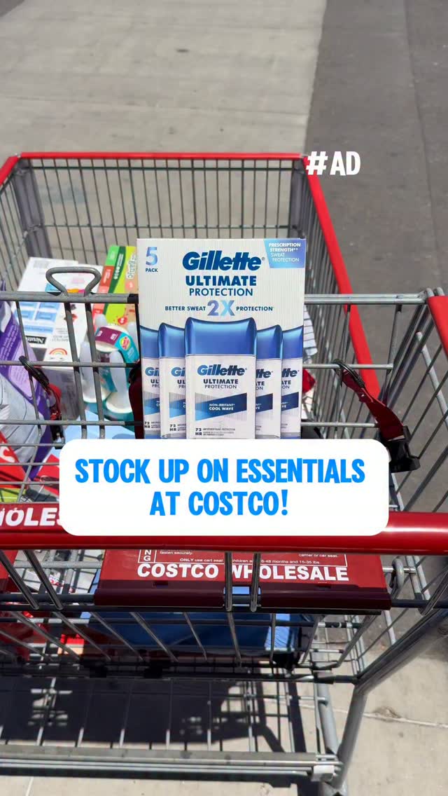 A shopping cart filled with various products, prominently featuring a multi-pack of Gillette Ultimate Protection deodorant. A large, white text box with blue lettering overlays the lower portion of the image, displaying the message "STOCK UP ON ESSENTIALS AT COSTCO!". The background shows a blurred outdoor scene, possibly a parking lot. The hashtag "#AD" is visible in the upper right corner.