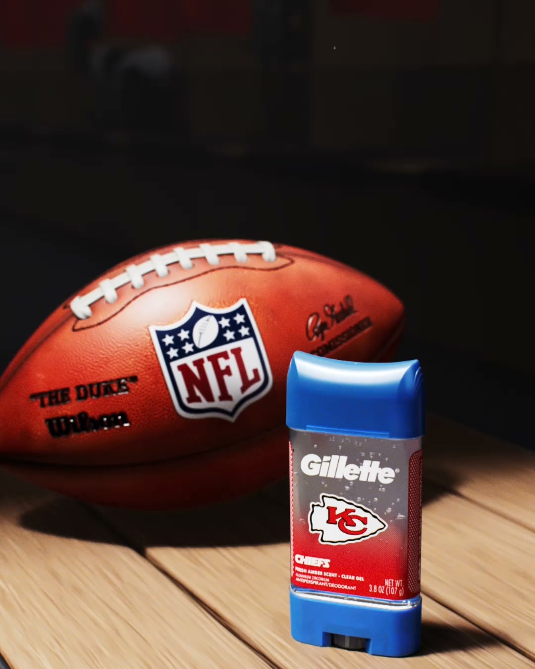 A Gillette deodorant stick with a Kansas City Chiefs logo is placed on a wooden surface next to an NFL football. The deodorant stick is blue and clear, with the Chiefs logo prominently displayed on its label. The football is orange and has the NFL logo and "The Duke" branding. The background is dark and out of focus, highlighting the product and the football. The on-screen text reads: "Get The Kingdom's Best Defense with Limited Edition Gillette Official Kansas City Chiefs Deodorant."