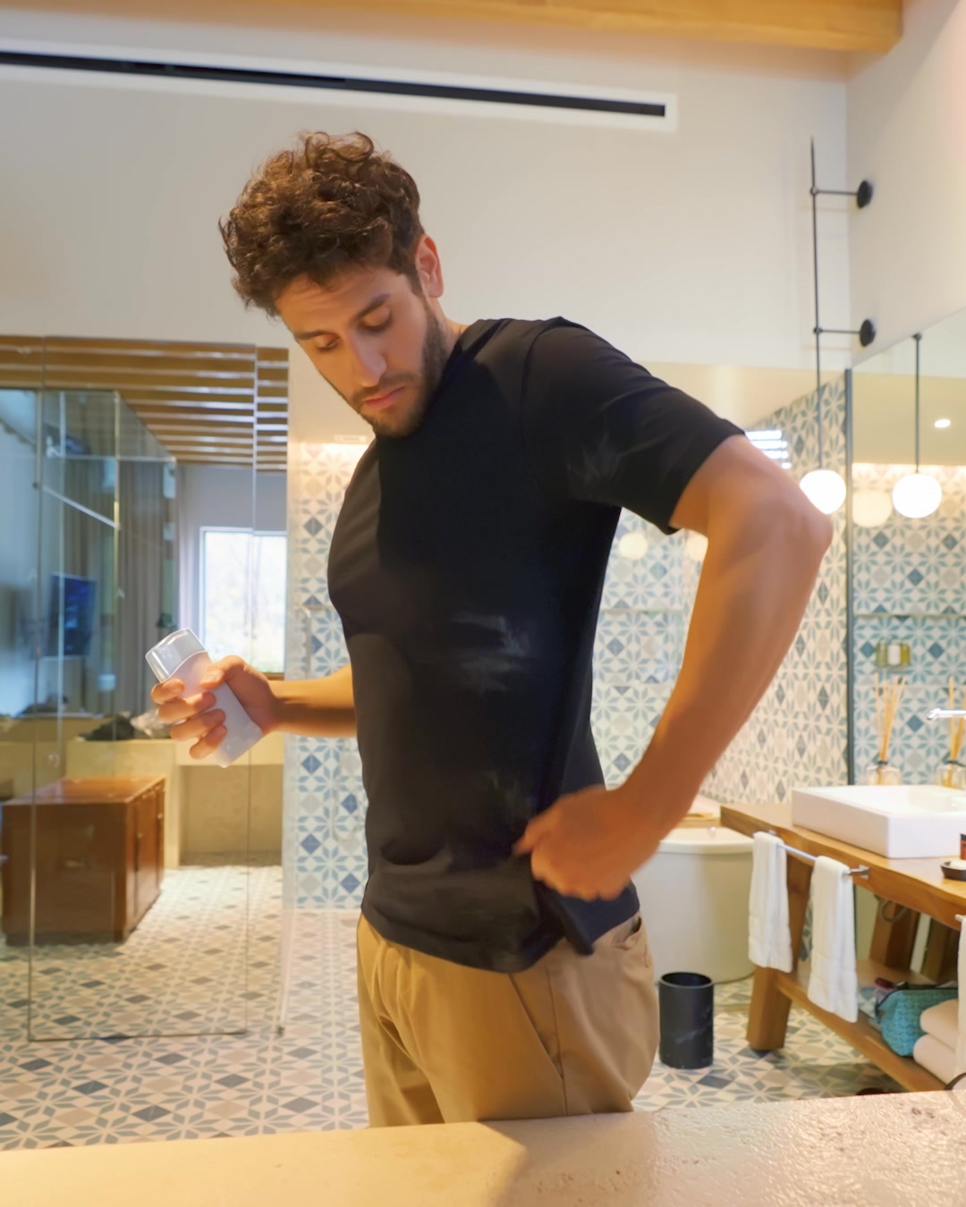 A man in a black t-shirt and tan pants stands in a bathroom, holding a white deodorant stick in his left hand and pulling up the side of his shirt with his right hand to reveal sweat marks. The bathroom has patterned tiles on the walls and floor, a sink, and a mirror. The text overlay states that Gillette is the #1 clinical brand for men with 2X better sweat protection compared to the moisture protection needed in a common antiperspirant.
